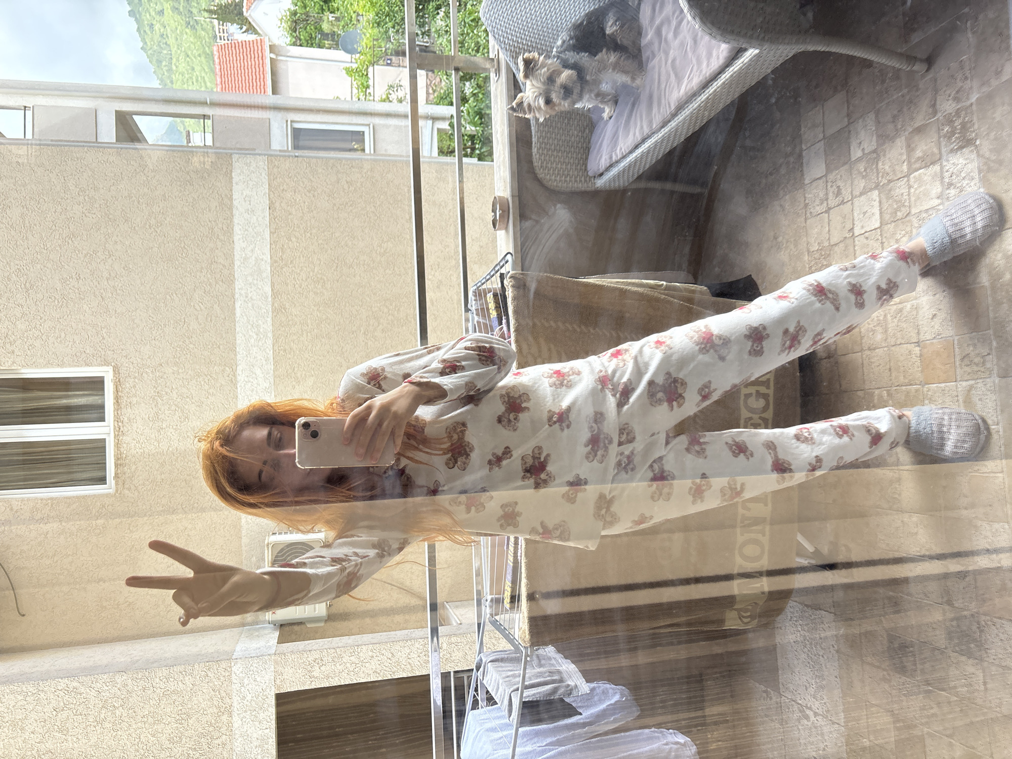A reflective image shows a woman in patterned pajamas and slippers, posing with a peace sign while taking a selfie on a balcony. A small dog sits on a wicker chair on the balcony. The reflection reveals an exterior view of buildings, possibly located in Budva, Montenegro, indicated by architectural styles and vegetation visible in the reflection. A clothing rack is on the balcony. The reflection shows a tiled floor.