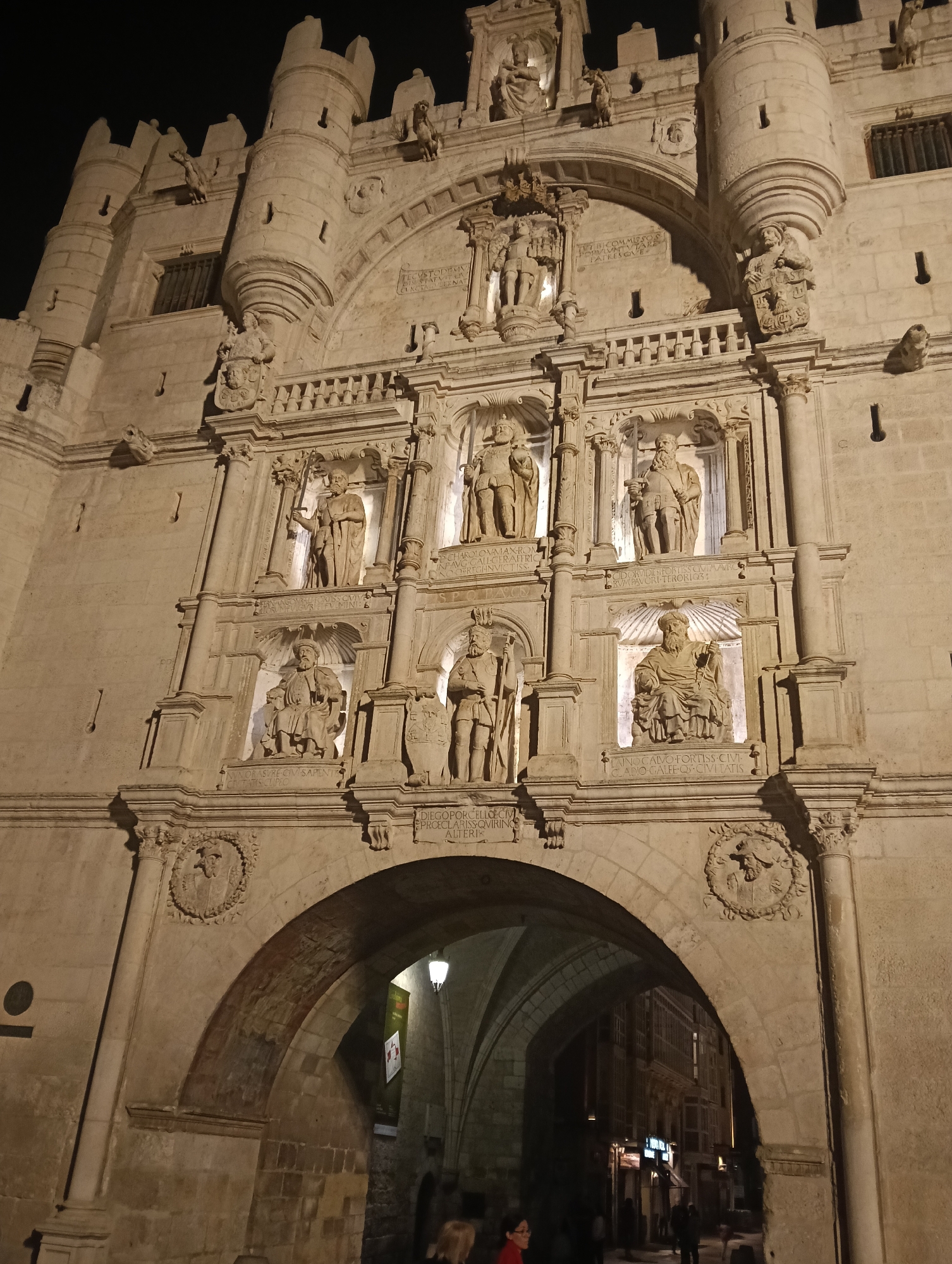 This image shows the Santa Maria Arch, one of the twelve medieval gates of the city of Burgos, Spain, at night. The arch is made of limestone and features several tiers of decorative sculpture, including figures in alcoves. A main archway leads through the structure into the city, revealing a glimpse of the street and buildings beyond. Some text is also visible on the arch's facade. The lighting illuminates the arch and its sculptures, contrasting against the dark night sky.