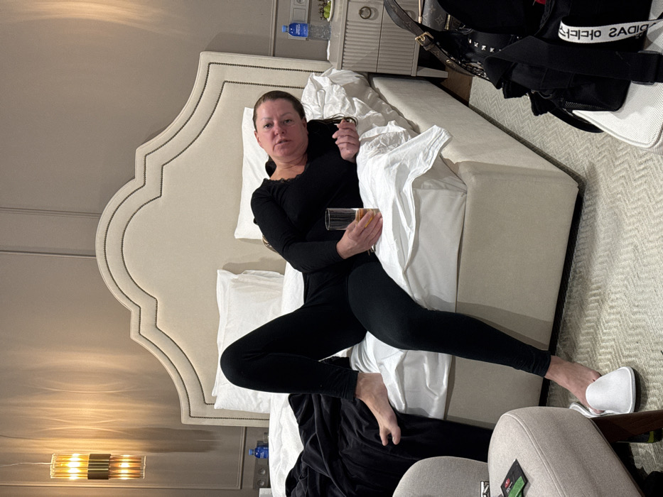 A woman in black leggings and a black long-sleeved shirt reclines on a bed with a large, ornate headboard. She holds a glass of orange liquid. The bed is covered in white linens. Her left foot is bare, while her right wears a white slipper. A nightstand sits beside the bed with a water bottle and other items. A jacket and a shopping bag are on the bed, and a chair is positioned at the bed's foot.