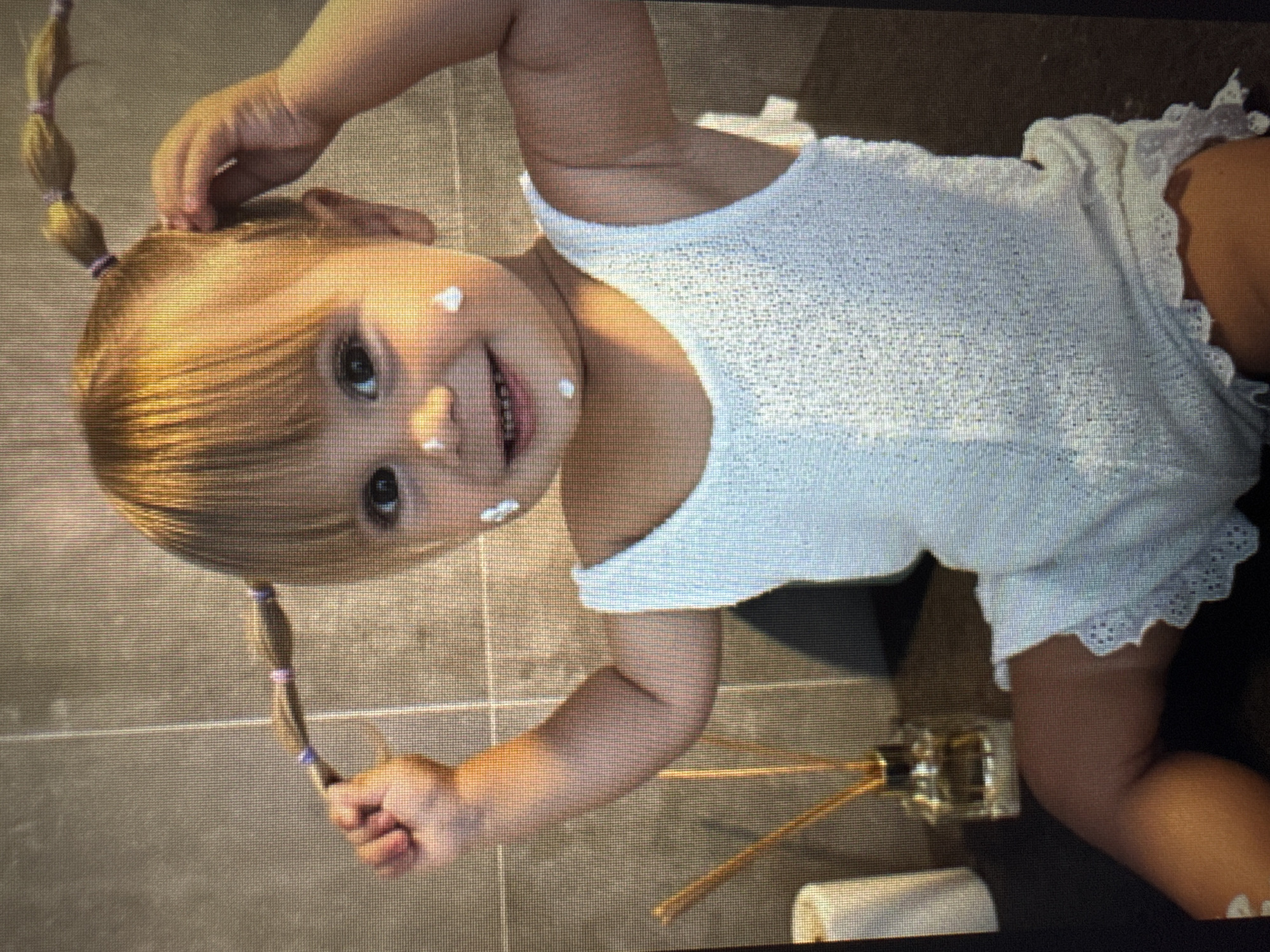 A young toddler with light blond hair and blue eyes smiles playfully at the camera. The child is wearing a white sleeveless top or dress made of a textured knit fabric with delicate lace trim along the hem. Their hair is styled into two pigtails, secured with purple elastic bands, and they have bangs across their forehead. Several white smudges of cream or lotion are visible on the child's face, specifically on the forehead, right cheek, and chin, suggesting playful exploration. One of the child's hands is raised to touch their hair near the top of the head, while the other hand holds the end of one of the pigtails. The child's mouth is open in a wide smile, showing teeth, and their eyes are looking directly forward. The setting appears to be indoors, with a floor composed of large greyish-brown square tiles visible in the background. A decorative reed diffuser with wooden sticks in a glass container and a folded white towel are also on the floor behind the child. The overall scene captures a moment of happy, mischievous innocence in a home environment. No visible text is present.