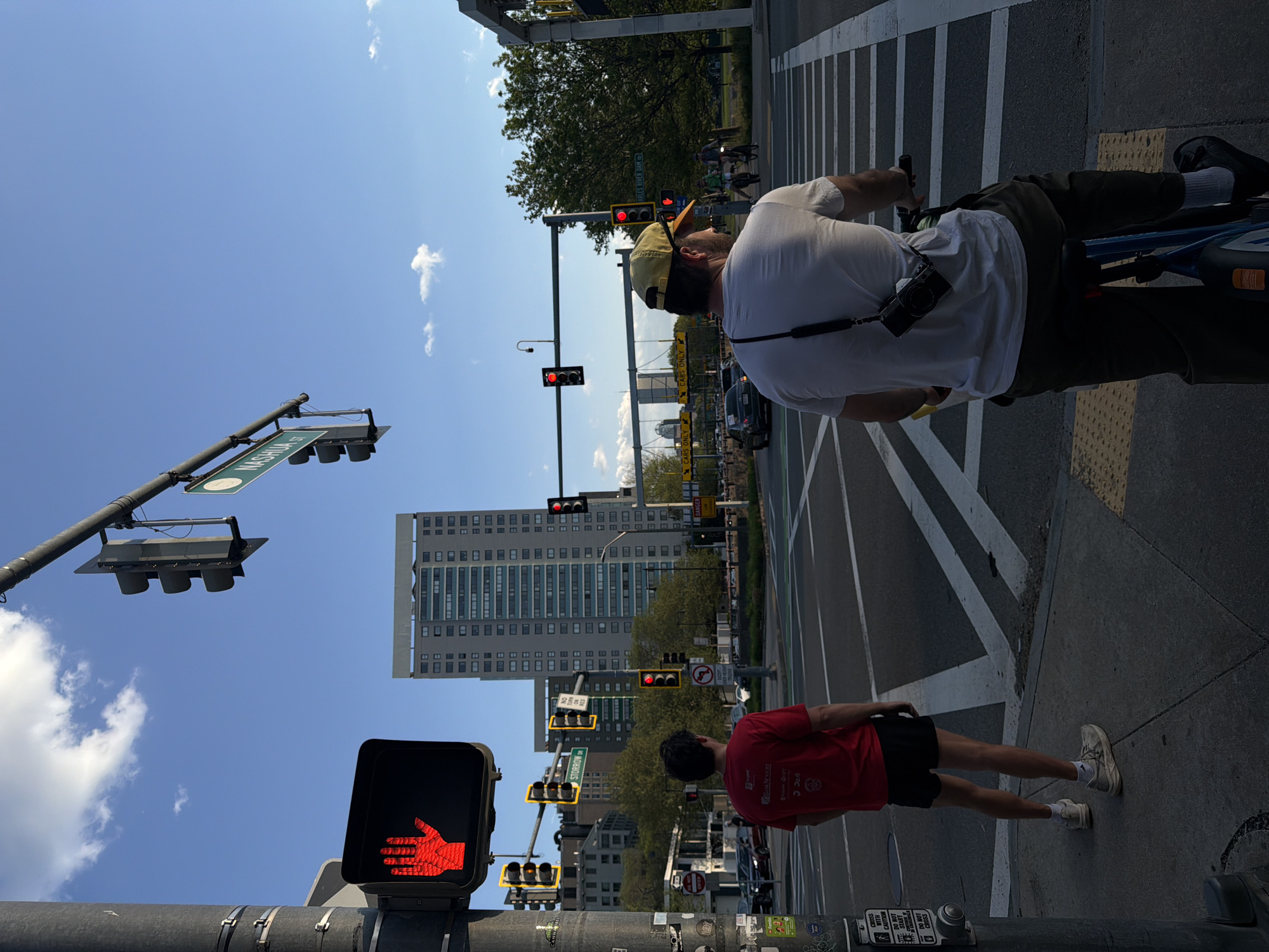 The image captures a city scene in Boston, Massachusetts, United States of America. It depicts two individuals waiting at an intersection controlled by traffic lights. One person, wearing a white t-shirt, yellow hat, and dark trousers, is on a bicycle. A black camera is slung around his neck. The second person, wearing a red t-shirt and black shorts, is standing on the sidewalk. All traffic lights visible in the frame are displaying red signals. Street signage indicates the intersection of Nashua Street and Storrow Drive. Several buildings are visible in the background, with one being a large, multi-story office building. Cars are visible on the street, and the sky is clear with a few scattered clouds. The image is rotated, with the sky towards the left.
