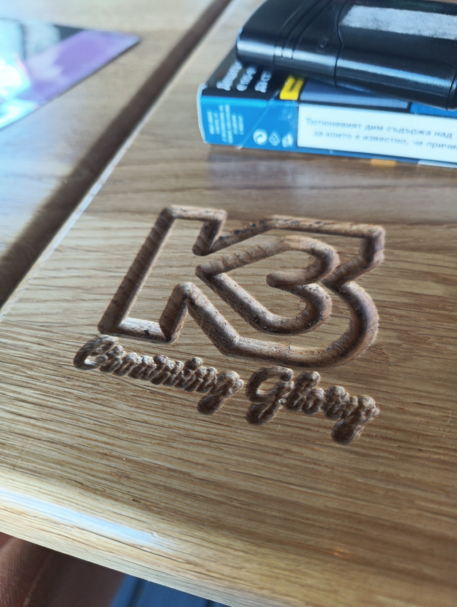 The image shows a light brown wooden surface with a carved design. The design consists of the letters "LB" in a stylized, interconnected manner, and the words "Burning Glory" written in a cursive font below. To the right and above the carved design, there is a blue and white box with text in a Cyrillic alphabet and a black object on top of it. The location is unknown, and Burgas, Bulgaria, cannot be determined from this image.