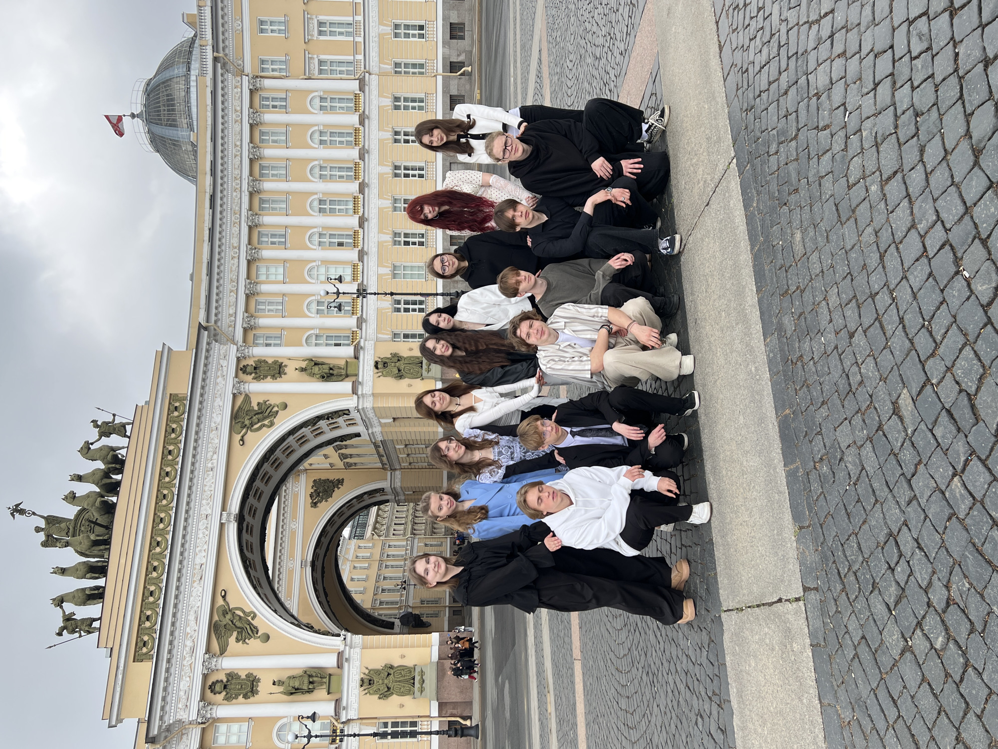 A group of about 15 people are arranged in rows, sitting on a paved area in front of a large, ornate, yellow building with a triumphal arch. The building has classical architectural elements, including columns, sculptures, and a quadriga on top of the arch. The people are a mix of genders and ages, wearing a variety of clothing styles. The ground is paved with dark grey cobblestones. The sky is overcast. The building appears to be part of the Winter Palace complex in Saint Petersburg, Russia, specifically facing Palace Square.