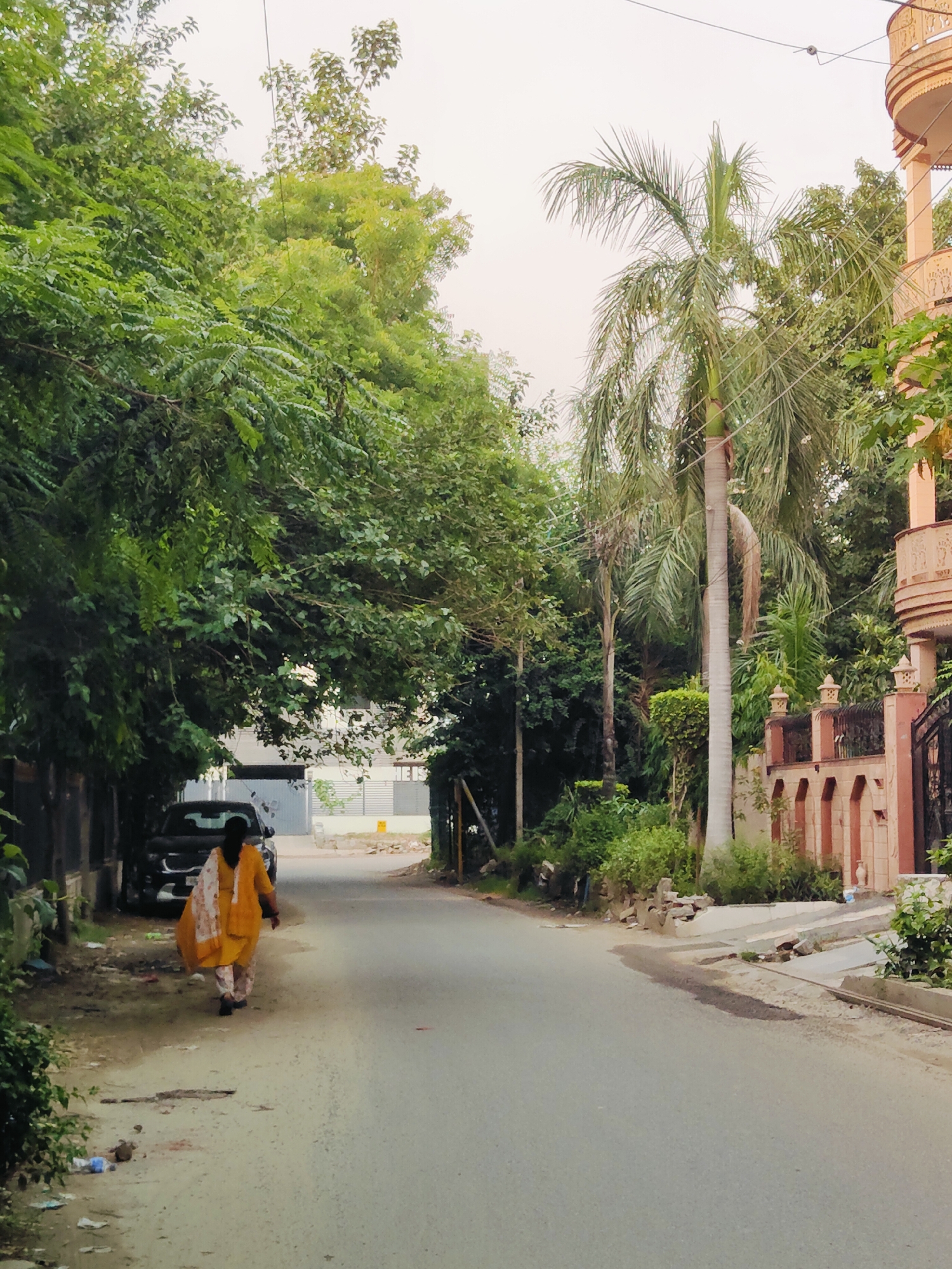 A woman in a yellow tunic and patterned pants walks away from the camera down a paved street lined with trees. To her left, a dark-colored SUV is parked. Beyond the trees on the left, a building with windows is visible. On the right side of the street, a decorative wall with arched openings, a gate, and a balcony of a building are visible. A tall palm tree stands prominently in the background. The sky is overcast and pale. The street is in a city, likely Gurgaon, India, given the architectural style and vegetation. The area appears to be a residential or mixed-use neighborhood with lush greenery and substantial buildings. The road surface is paved, and the edges are lined with dirt and some debris.