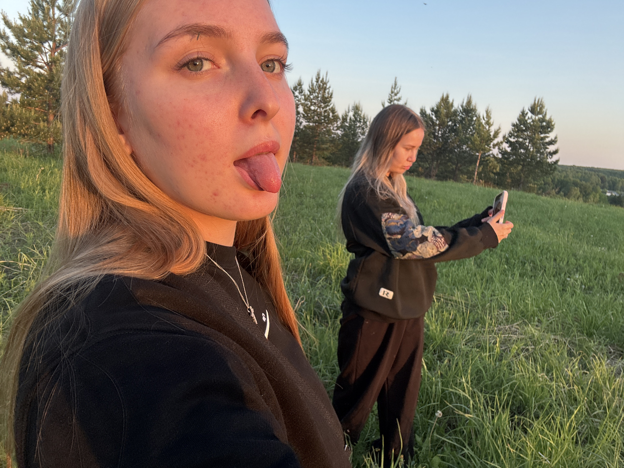 Two young women are standing in a grassy field. The woman in the foreground is close to the camera and has blonde hair, light skin, and is sticking her tongue out. She is wearing a black sweatshirt with a silver necklace. The other woman is standing further back, holding a cell phone and appears to be taking a picture. She has blonde hair, is wearing a black sweatshirt with floral print sleeves and black pants. The background includes trees on a hill and a clear sky. The location is Zhigalki, Russia.