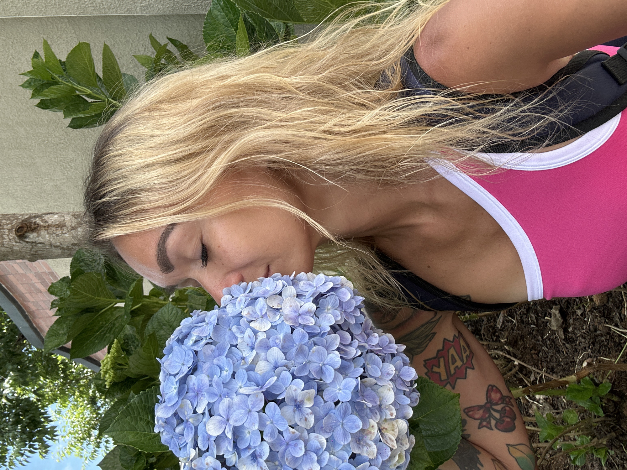 In Da Lat, Vietnam, a woman with blonde hair and visible tattoos on her arm is seen smelling a large cluster of blue hydrangeas. She is wearing a pink tank top with a white trim and a dark backpack. The background shows lush green foliage, a tree trunk, and a brick wall.