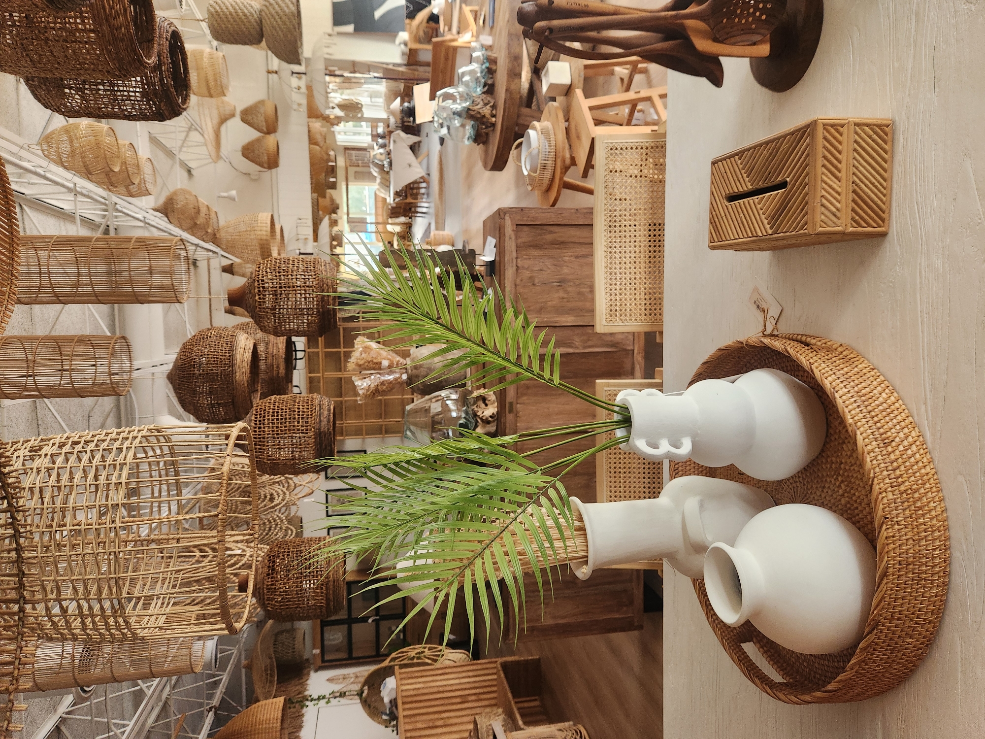 The image shows a retail store interior with a focus on home decor items. In the foreground, a white textured surface displays a woven circular tray holding three white ceramic vases of varying shapes and sizes. To the right of the tray is a rectangular woven bamboo box with a slot on top, likely a tissue box holder or decorative storage. Further to the right, a dark wooden utensil holder with several wooden spoons is visible. Behind these foreground objects, a display of green palm fronds in white vases adds a touch of natural greenery.
The mid-ground reveals more of the store's offerings, including wooden furniture such as cabinets and tables, chairs with woven cane seats and backs, and more decorative items like glass spheres on wooden bases. The background shows a broader view of the store, with more tables, chairs, and hanging lamps made of natural materials like rattan and bamboo, creating a bohemian or coastal aesthetic. The store appears to be well-lit, with natural light possibly coming from windows or skylights in the ceiling. The overall impression is a curated retail environment showcasing furniture and decor.
The image was taken in Maitland, United States of America, although a specific address or business name is not provided.