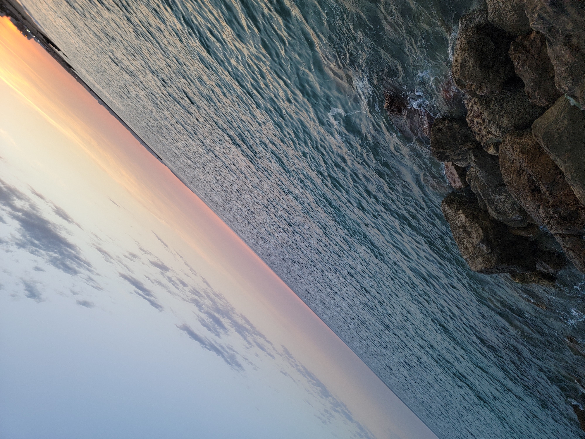 The image depicts a diagonal view of the Atlantic Ocean off the coast of Quarteira, Portugal, during what appears to be sunset or sunrise. The sky transitions from a soft blue on the left to pastel pinks and oranges on the right, with scattered clouds. The ocean is a deep teal color, with visible ripples and waves that catch the light. Along the bottom right of the frame, a rocky shoreline is visible, with dark, textured rocks interspersed with small waves lapping at their base. In the distance, along the horizon where the sky meets the water, a faint line of buildings and structures suggests the presence of the Quarteira coastline. There are no people or discernable actions taking place in the scene; it is a static landscape view.