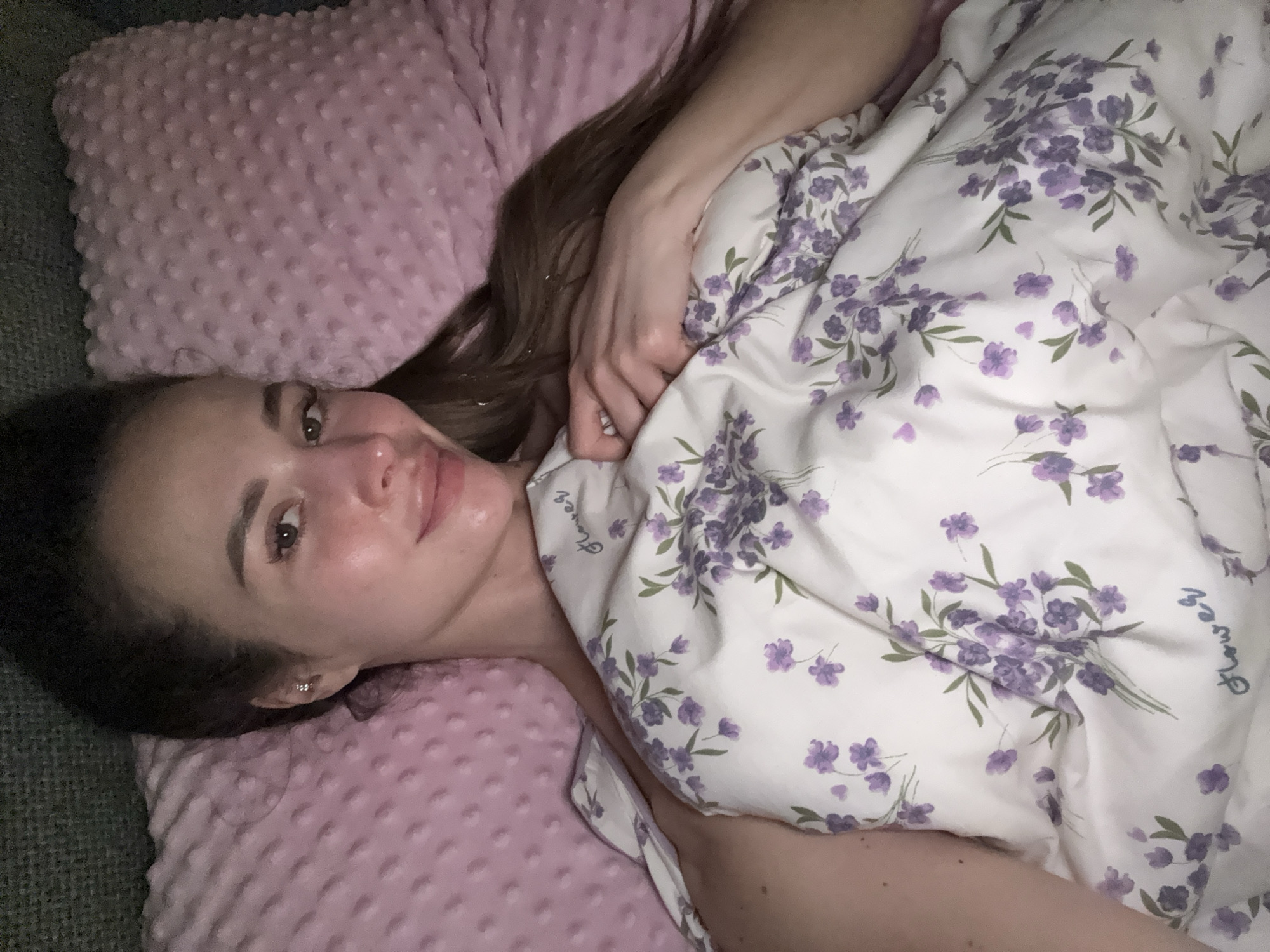 An indoor, portrait-oriented shot captures a young woman lying down, looking directly at the camera with a soft, pleasant expression and a gentle smile. Her head rests on a light pink, textured pillow, possibly made of minky fabric with raised dots. Her long, dark brown hair spreads out behind her head and shoulders. She wears a small, shiny earring in her left ear.
She is partially covered by a white duvet or blanket adorned with a delicate lavender floral pattern featuring scattered purple flowers and small bouquets with green leaves. The word "Flower" is subtly visible in cursive script on the duvet. Her left hand is visible, resting on the blanket. The background to her left appears to be a dark, textured surface, possibly a couch or another piece of bedding.
The setting appears cozy and relaxed, suggestive of a peaceful moment. The lighting is soft and even, typical of an indoor environment. The overall mood is calm and comfortable. This scene takes place in Kyiv, Ukraine.