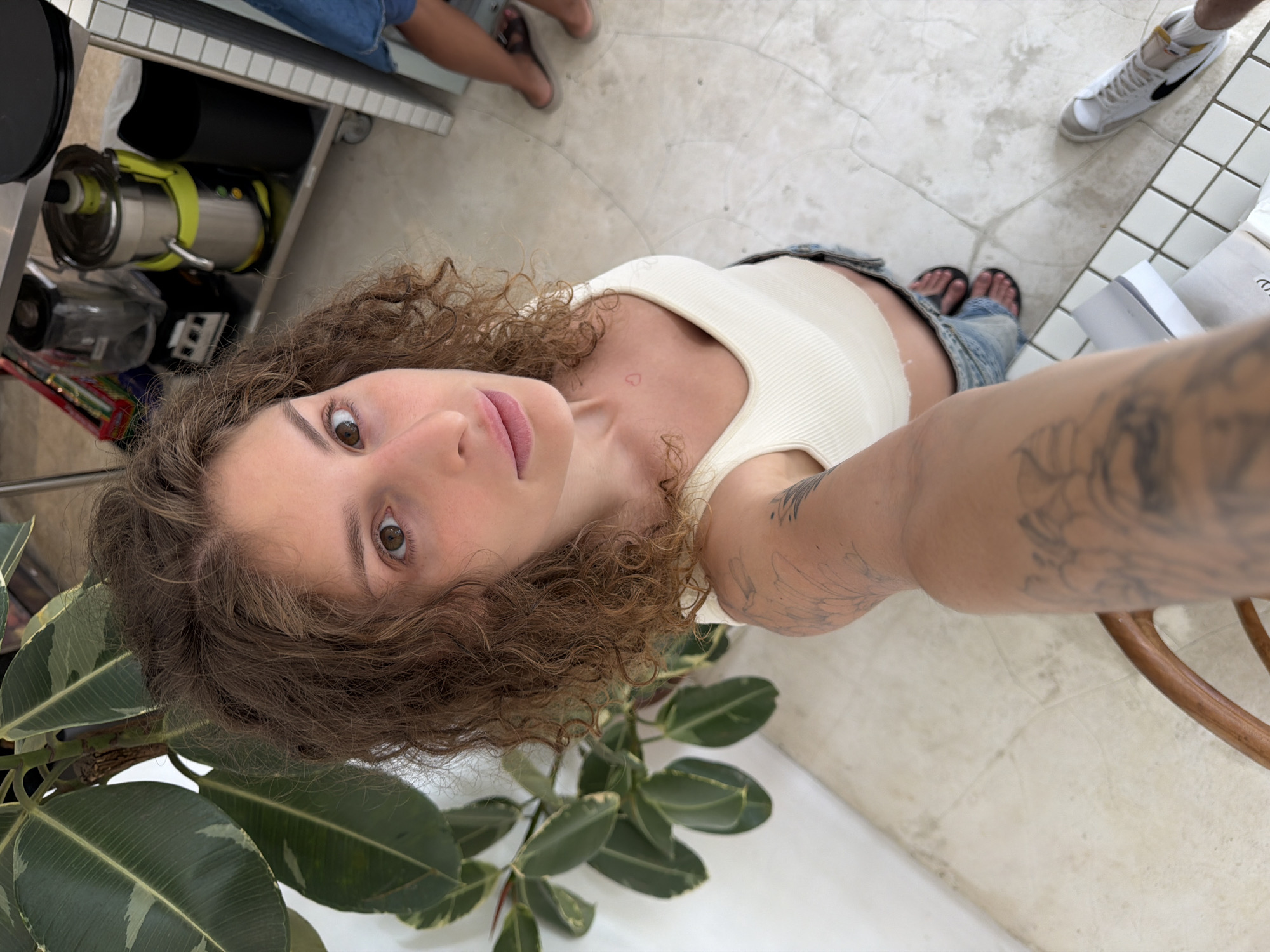 In an indoor setting, potentially a cafe or shop, a young woman with curly brown hair takes a selfie from a high angle. She is wearing a white ribbed tank top and denim shorts. A small, faint red heart tattoo is visible on her chest. Her left arm, which is holding the camera, is covered in tattoos. To her left, a large potted plant with broad green and yellow variegated leaves is prominent. In the background, a stainless steel cart holds various kitchen appliances, including a blender and a juicer. The floor is tiled with light-colored square tiles. Another person's legs, wearing denim shorts and dark sandals, are visible on the right side of the frame. White sneakers are also present on the floor. The overall lighting is bright, suggesting daylight. The location is identified as Mengwi, Indonesia.