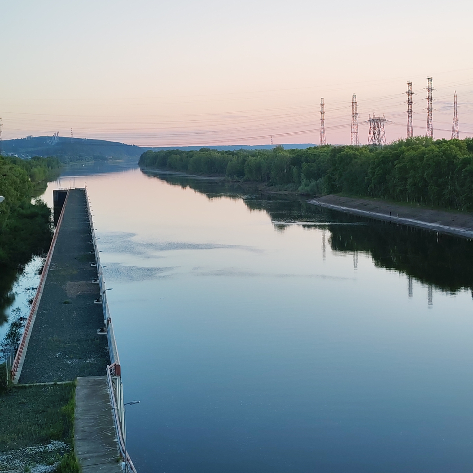 The image captures a wide, tranquil river scene at either dawn or dusk, with a sky transitioning from soft pink to light blue hues. The water, appearing calm and still, perfectly reflects the sky and the surrounding banks. On the left, a long, dark-colored concrete and gravel structure, resembling a pier or part of a lock/dam system, extends into the distance, bordered by a railing and featuring small lights or bollards along its edge.
Both sides of the river are lined with dense green forests. The right bank features a distinctive engineered embankment where two small, indistinct human figures can be observed walking or standing near the water's edge. Dominating the distant skyline are numerous power transmission towers, varying in design and height, some with complex lattice structures, silhouetted against the pale sky and running along the crest of a low hill. Further structures are faintly visible on the distant hillsides, integrated into the landscape.
The scene combines natural beauty with significant industrial infrastructure. The time of day suggests a peaceful atmosphere. No visible text is present in the image. The location is Chaikovsky, Russia.