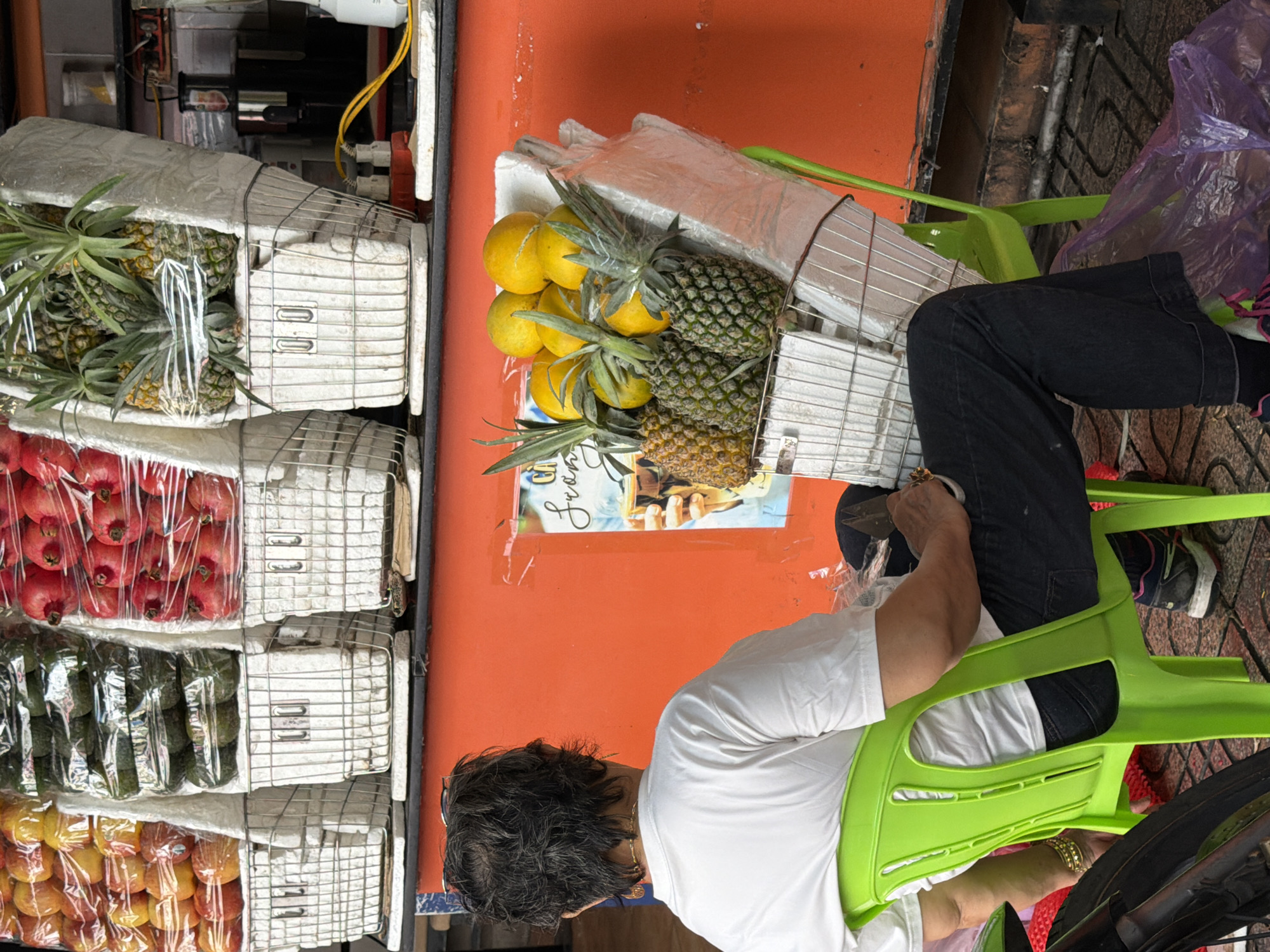 The image captures a street vendor scene in Nha Trang, Vietnam. A person is seated on a bright green plastic chair, facing away from the camera. They are wearing a white t-shirt, black pants, and sneakers. The individual appears to be a vendor, as they are positioned behind a display of fresh fruits.
In the background, a stall is stocked with various fruits, including pomegranates, avocados, pineapples, and oranges, all displayed in white wire baskets. The fruits are partially wrapped in plastic. Behind the fruit display, an orange wall serves as the backdrop, with some posters and electrical outlets visible. The ground is paved with patterned tiles. A motorcycle is partially visible on the left side of the frame. The overall scene suggests a typical street-side fruit stand operating in an urban environment.