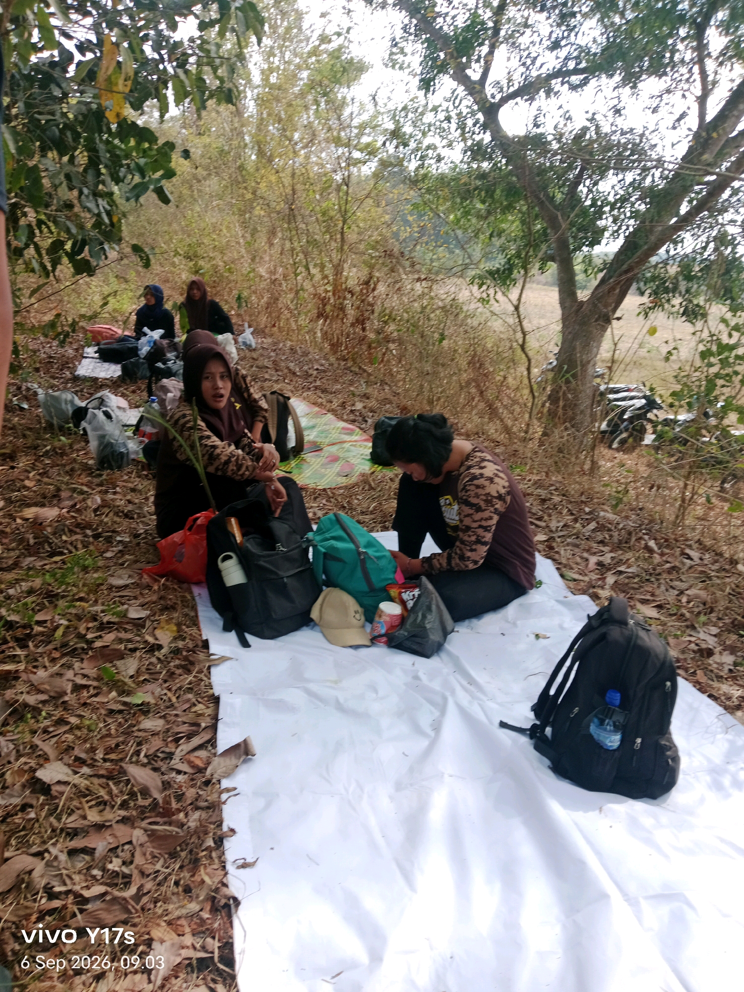 In an outdoor, wooded area with dry leaves on the ground, four individuals are seated on white and patterned mats. Three women, two wearing headscarves and one with short dark hair, are visible. The woman in the foreground wearing a headscarf is looking towards the camera with an open mouth, and appears to be speaking. She is wearing a camouflage patterned top. The second woman, also wearing a headscarf, is seated to her left. The third woman, with short dark hair, is seated to the right of the first woman, looking down at her hands. To the far left, partially visible, is another individual. Scattered around them are several bags, including black backpacks, a green backpack, a red bag, and other assorted bags and items. In the background, there are trees with dry branches and leaves, and a hint of a dirt path. To the far right of the image, several motorcycles are parked. The scene depicts a gathering or rest stop in a natural setting. The timestamp on the bottom left corner indicates "vivo Y17s 6 Sep 2026, 09.03".