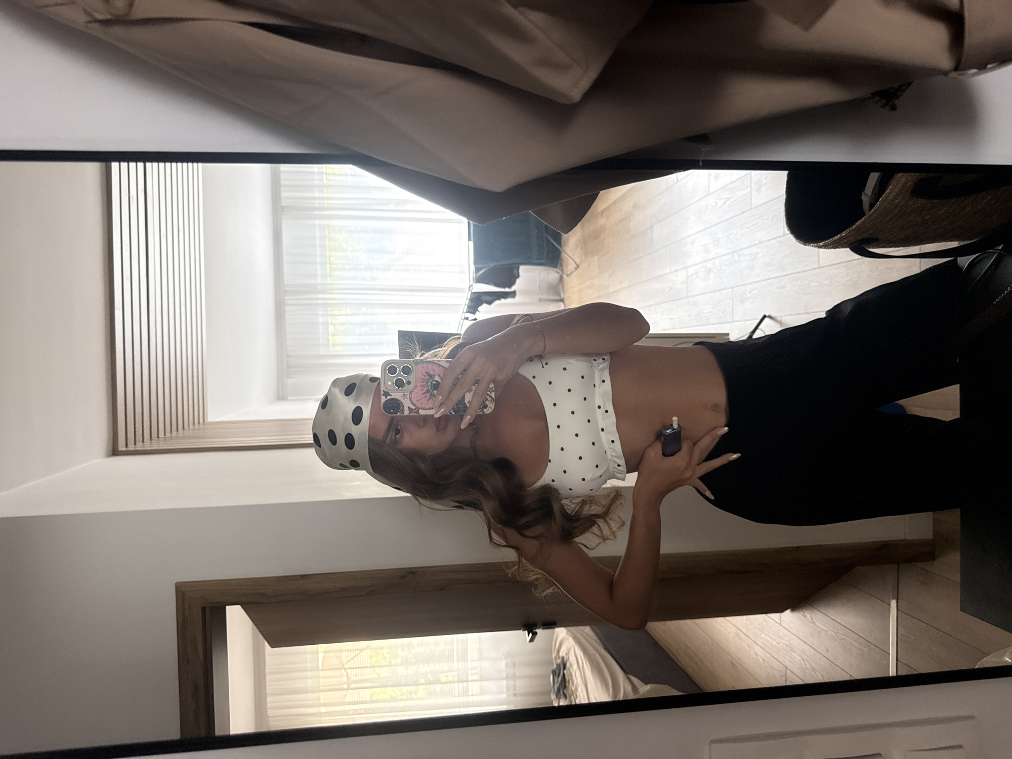 A young woman with long, wavy brown hair, wearing a white polka dot bikini top and black pants, stands in front of a mirror and takes a selfie. She is also wearing a polka dot headscarf. In her right hand, she holds a mobile phone, and in her left hand, she holds a small dark object. The background shows a bedroom with a bed, a wooden door, and a window with white curtains. A tan jacket hangs on the right side of the mirror. There is no indication of the city of Plovdiv, Bulgaria.