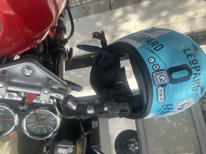 A turquoise "LEOPARD RACING" motorcycle helmet is hanging by its chin strap from the left handlebar of a red Suzuki motorcycle in Ngũ Hành Sơn Ward, Vietnam. The helmet is inverted, displaying "LEOPARD RACING" text, markings "A138-79", "QUATEST 2", "QC", and size "L" stickers. The motorcycle features chrome handlebars, a black left-side control module, and a dashboard with two circular gauges, one visibly labeled "SUZUKI" with a speedometer indicating up to 140 km/h. A key with a red plastic fob is inserted into the ignition. A black phone holder labeled "Motorcycle/Bicycle" is mounted on the handlebars. The motorcycle is parked on a light-colored paved surface, with tree shadows visible. A blurred reflection in the background shows a building interior, including a circular object resembling a washing machine drum with the number "01" visible. No individuals are present.
