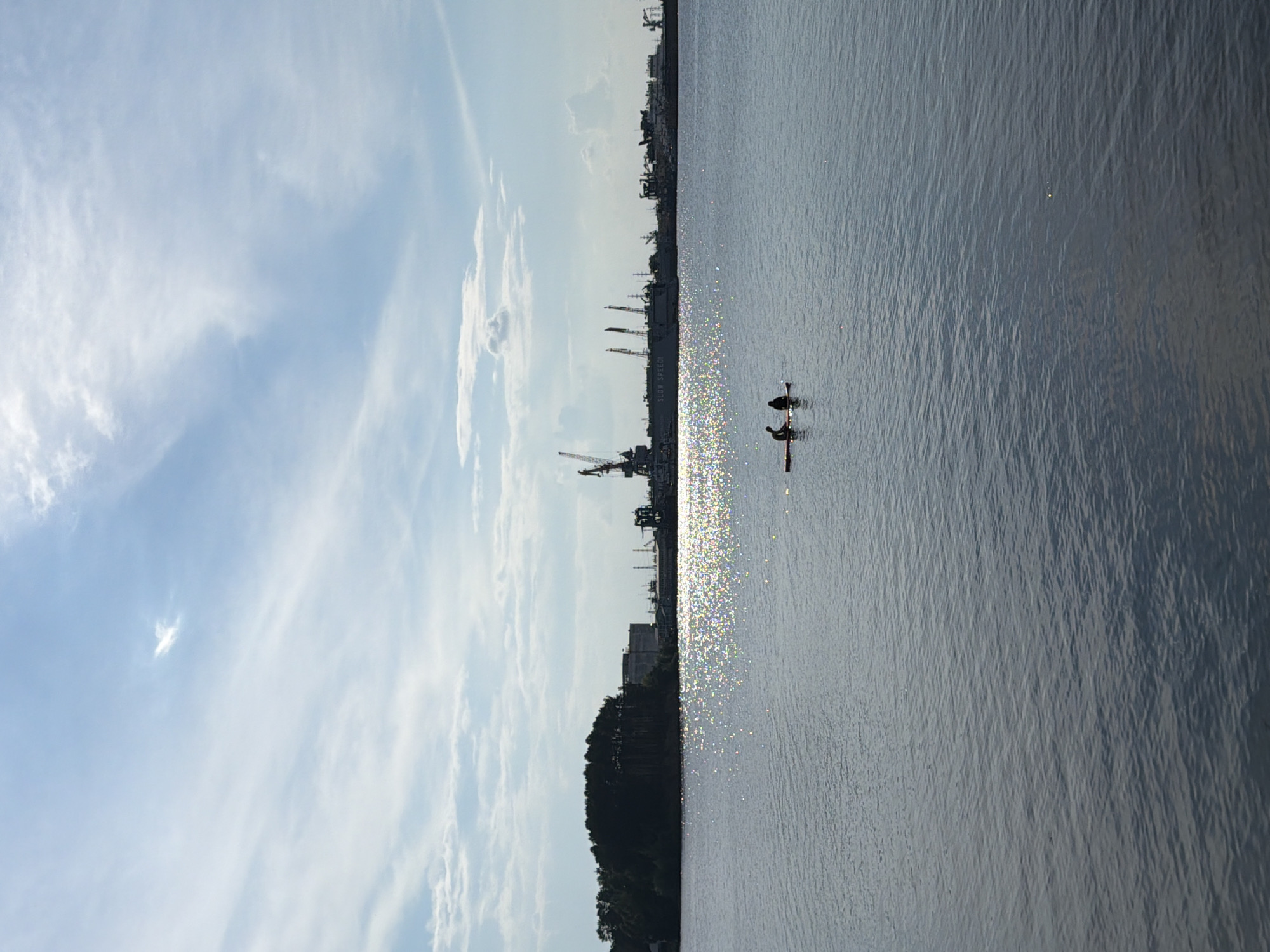 In Chaikovsky, Russia, the image captures a wide, calm body of water reflecting the bright blue sky dotted with white clouds. In the distance, a tree-covered landmass separates the water from a complex industrial harbor featuring numerous cranes and docked ships. Two silhouetted figures are visible in the middle of the water, paddling on a small raft. The sunlight creates a shimmering effect on the water's surface.