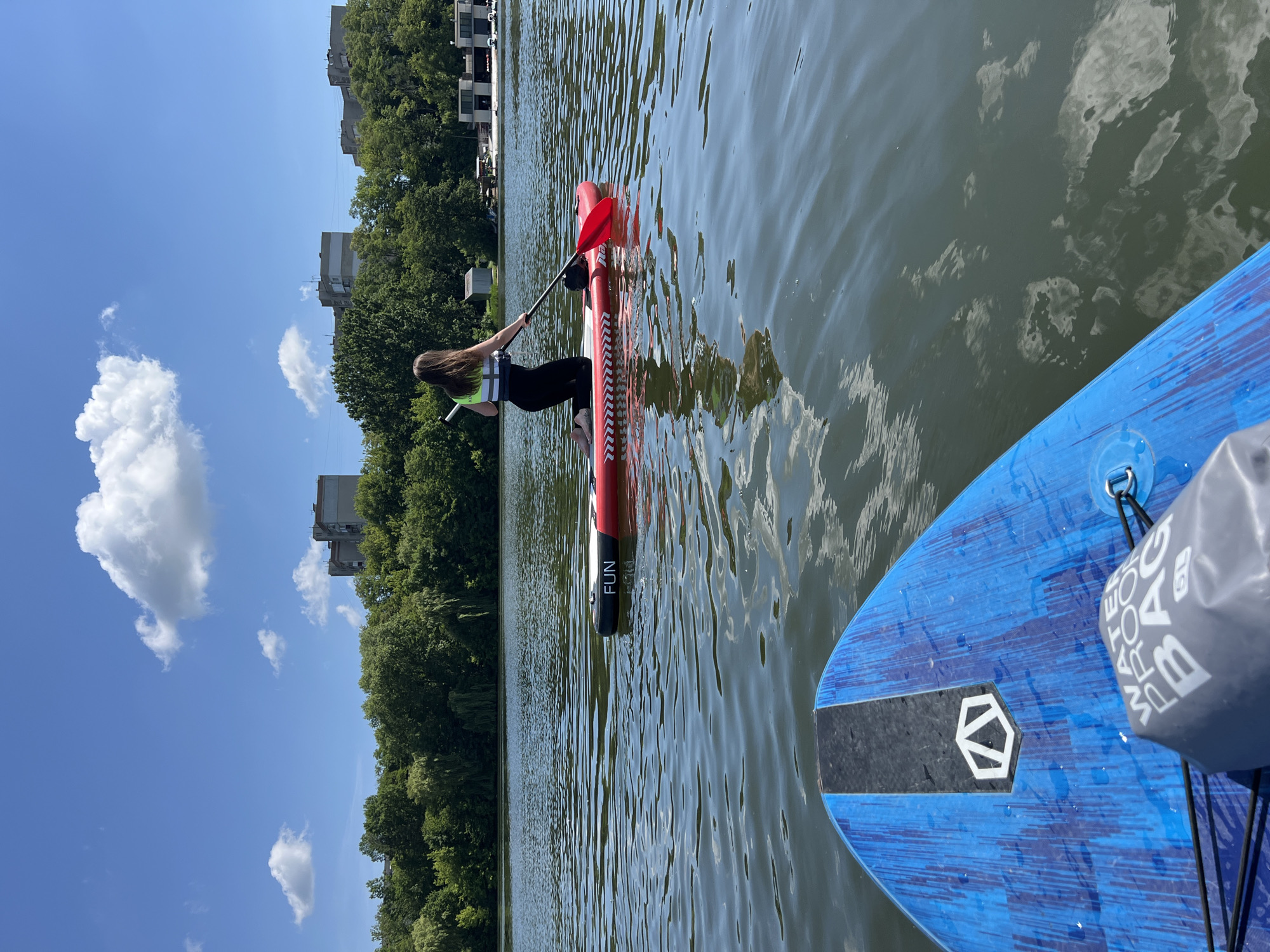 The image captures a sunny day on a body of water, likely Ternopil Lake, in Ternopil, Ukraine, as evidenced by the presence of apartment buildings and a lush green treeline in the background, typical of urban landscapes near the lake. In the foreground, the viewpoint is from a blue and black stand-up paddleboard. Further out on the water, a young woman with long brown hair is kneeling on a red and black stand-up paddleboard. She is wearing a white life vest over a dark top and dark leggings, and she is holding a red paddle, actively engaged in paddling. The water is calm with gentle ripples reflecting the blue sky and scattered white clouds. The shoreline is lined with dense trees, and beyond them, several multi-story apartment buildings are visible, indicating an urban setting.