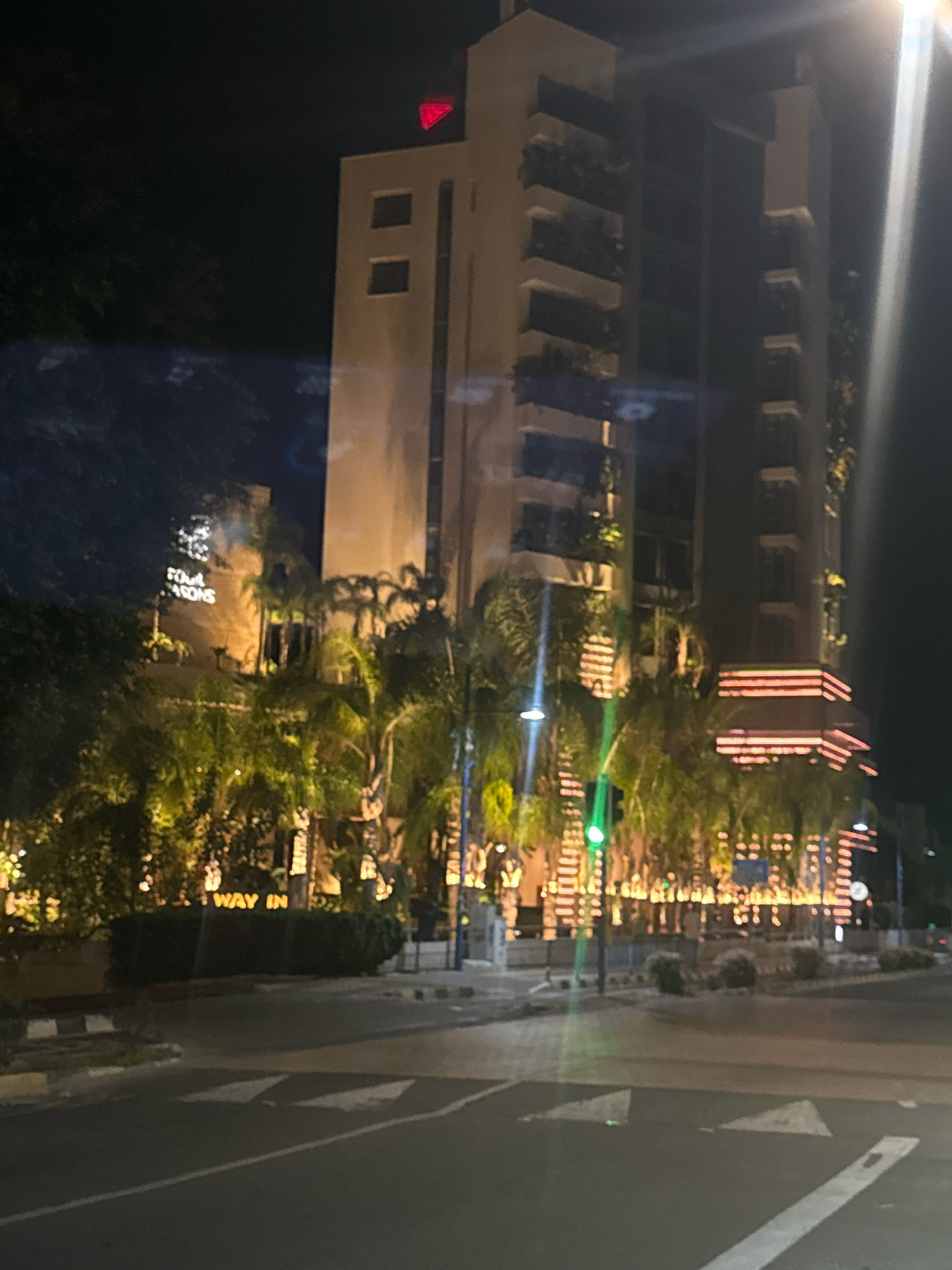 A nighttime, medium-shot vertical photograph captures the illuminated exterior of the Four Seasons Hotel in Agios Tychona Community, Cyprus. The hotel, a prominent building, is adorned with festive lighting that outlines its architectural features and surrounding palm trees. The facade is a light beige color, with darker sections and balconies. The hotel's signage, "Four Seasons," is visible and lit up. In the foreground, a street with traffic markings and a green traffic light is present, indicating a roadway. The scene suggests a vibrant and lively atmosphere, characteristic of a tourist destination.