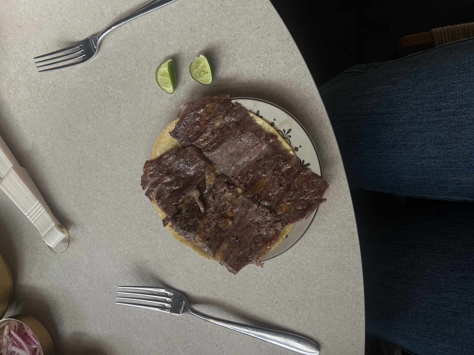 On a light gray tabletop are several objects. Centered is a small plate with a brownish tortilla topped with dark brown meat. To the left of the plate are two lime wedges. A fork is above the limes, and another fork is below the plate. In the bottom left corner, there is a container of pickled onions. To the right of the plate, denim fabric suggests the presence of a person. The location is Unknown, Unknown.
