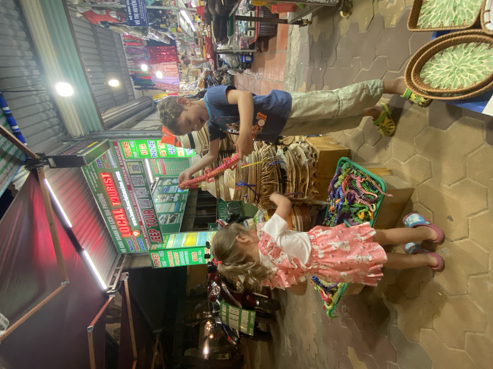 In Phu Thuy Ward, Vietnam, a boy and a girl stand by a stall selling woven hats and colorful braided bracelets. The boy, wearing a blue t-shirt and beige pants, holds a pink braided item. The girl, in a pink dress with rabbit prints, is reaching into a green bin filled with braided bracelets. Behind them, a sign advertises "LOCAL TOURIST" services, including Jeep Tours, and offers motorbike rentals. Scooters are parked to the left. The scene is illuminated by fluorescent lights and the red glow of an awning. The ground is paved with hexagonal tiles.