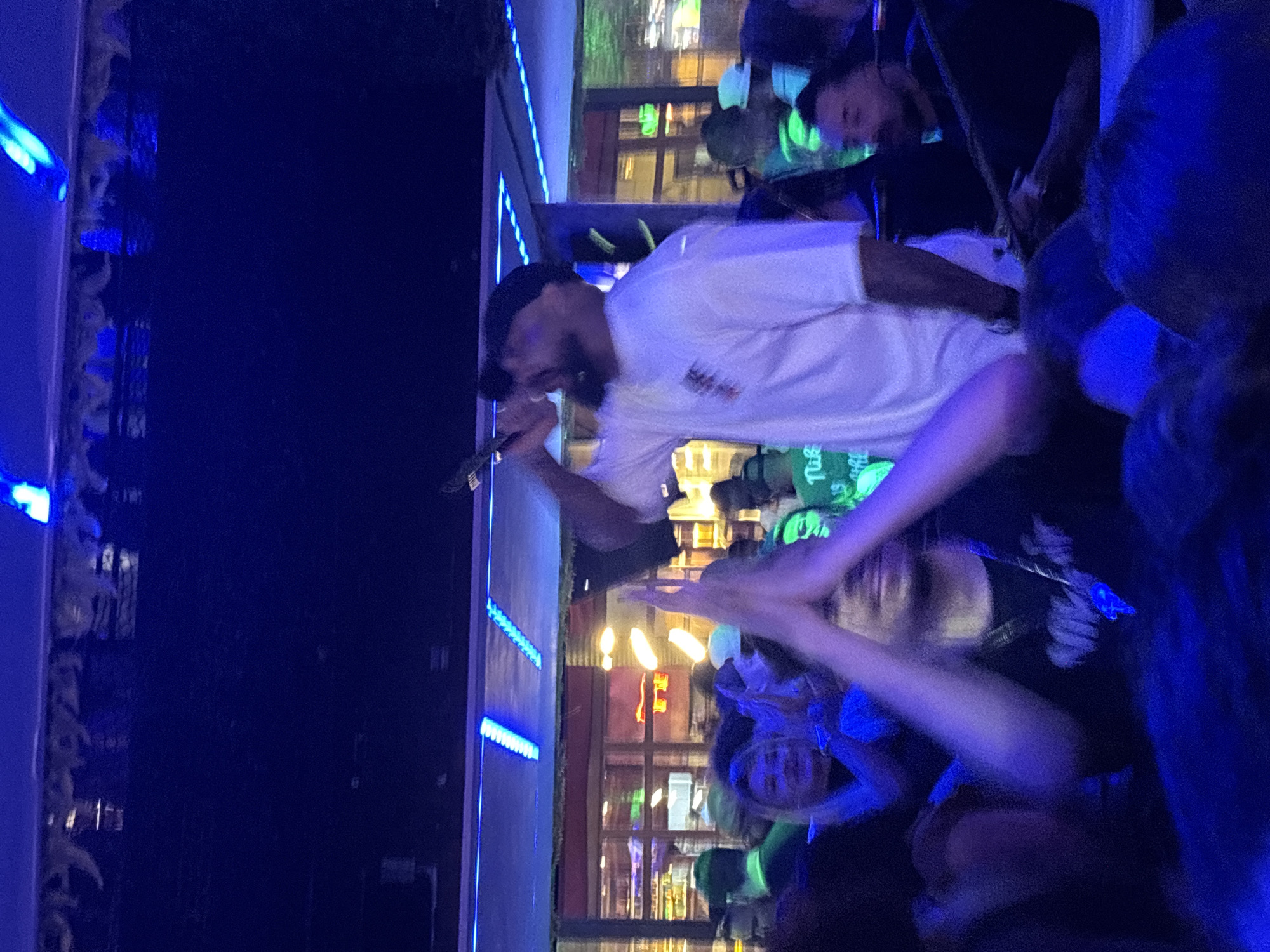The image depicts a live music performance. A male singer with a beard and a black baseball cap is holding a microphone, singing, and wearing a white t-shirt. The background suggests an urban environment with illuminated buildings, possibly a bar or restaurant with windows. There are audience members in the foreground and background, some with hands raised, presumably enjoying the performance. Blue lighting effects are visible. The location is likely a live music venue or outdoor event in an urban setting, possibly in São Paulo, Brazil, although visual clues within the image do not specifically pinpoint São Paulo.