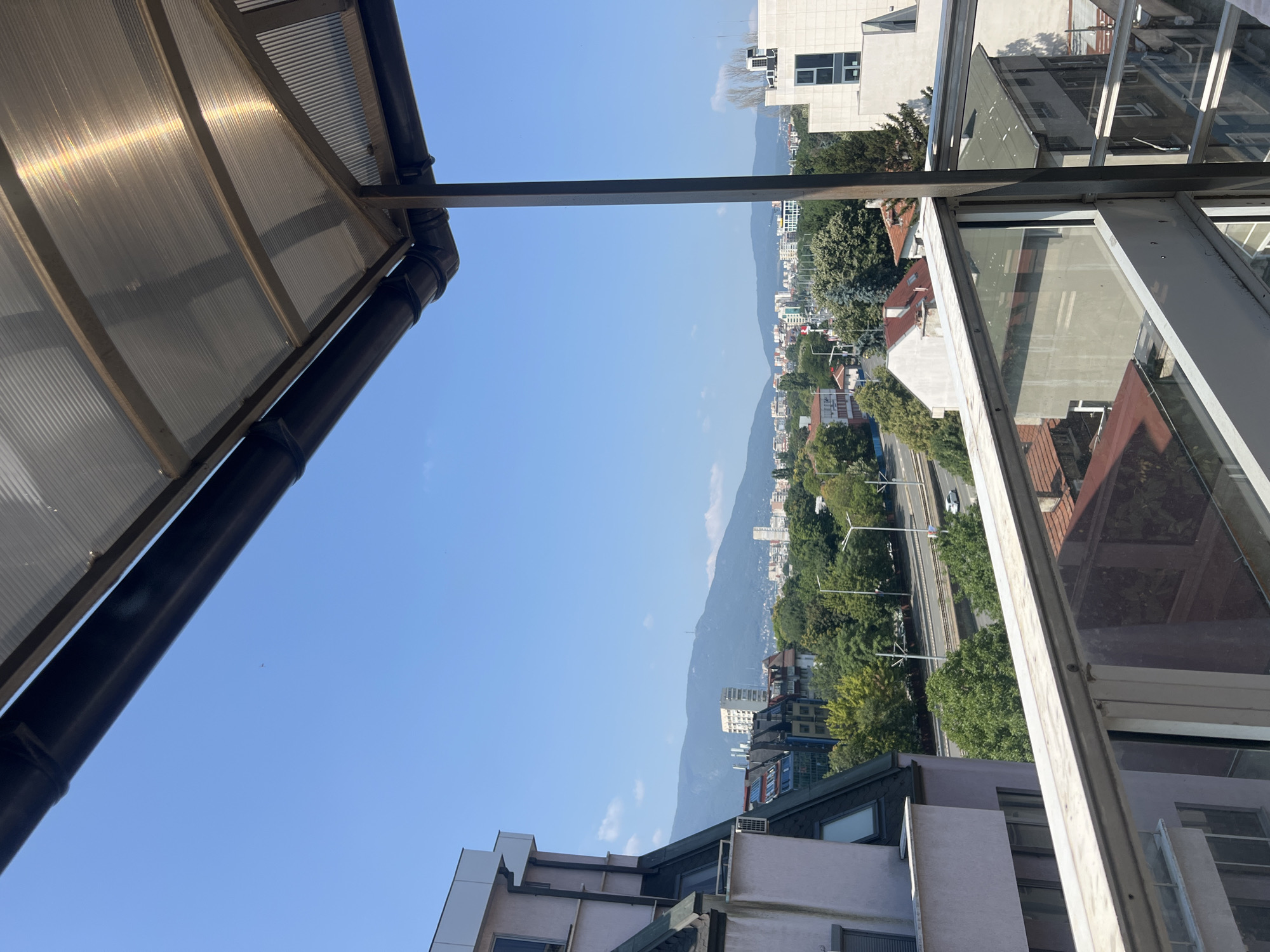 This is a wide-angle, eye-level shot of a cityscape, likely taken from a balcony in Sofia, Bulgaria. The foreground features a clear sky and a view of a modern building and a balcony railing. The midground showcases a busy street with vehicles, lush green trees, and various residential and commercial buildings. In the distance, a mountain range, possibly Vitosha Mountain, forms a picturesque backdrop, with a prominent telecommunication tower visible on one of the peaks. The overall atmosphere is bright and sunny, suggesting a clear day.