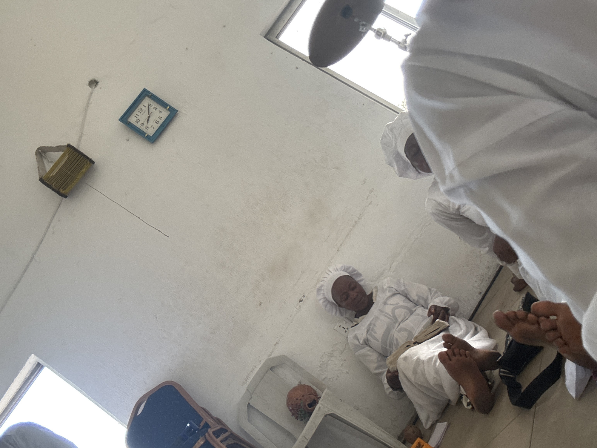 A medium shot depicts a room with a blue-framed clock and a yellow object hanging on the wall on the left side. A person is lying on the floor in white garments, while two other people in similar attire are seated nearby. Bare feet are visible in the lower right corner. A blue chair and a cabinet with an object inside are situated near the bottom of the image. The location is likely in the city of Badagry, Nigeria.