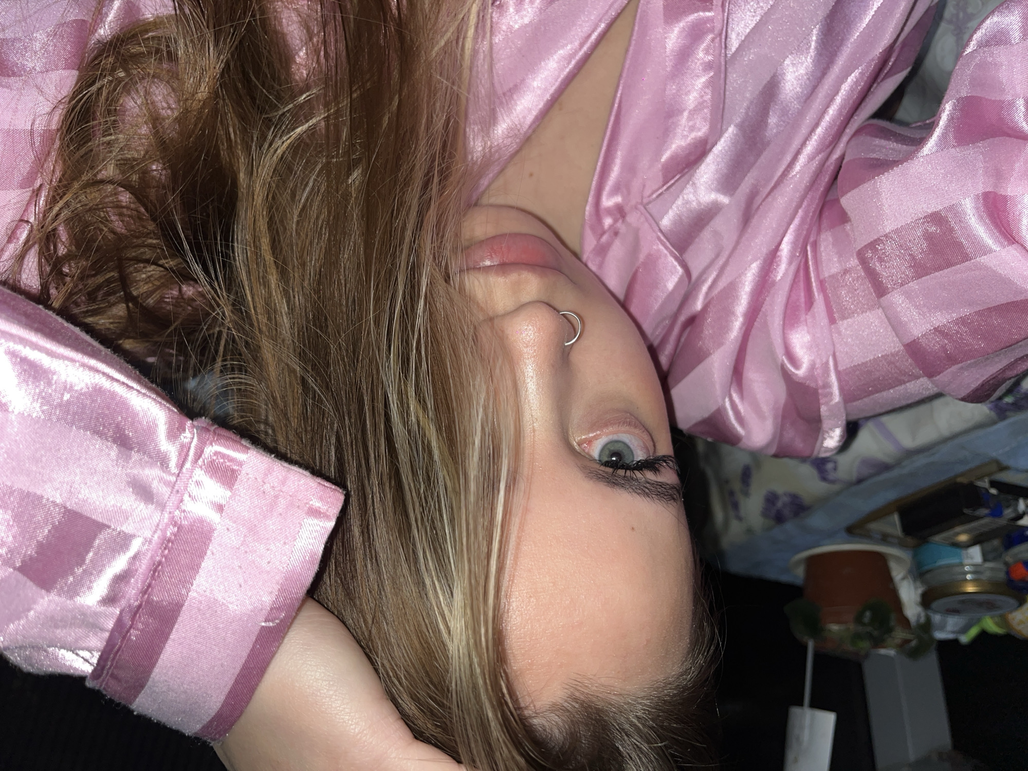 A person with blonde and brown hair is photographed from an unusual perspective. The person is wearing a pink striped pajama top. A silver nose ring is visible on the left nostril. The background is slightly blurred, revealing objects like a plant, jars, and a picture frame on a surface. The setting seems to be indoors, potentially a bedroom. There's no information provided to determine whether this image was taken in Novopokrovskaya, Russia.