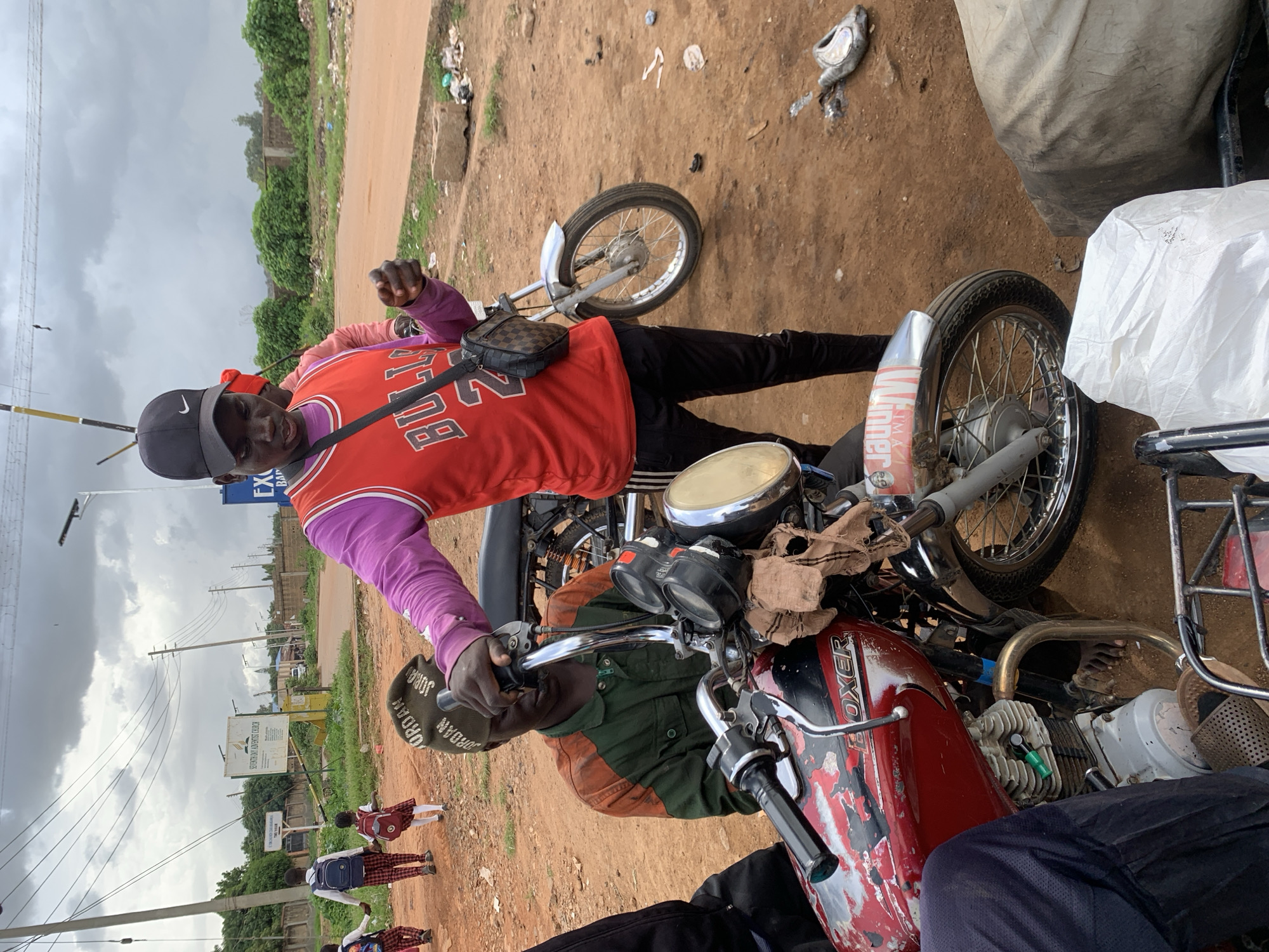 This media file captures a street scene in Barakin Lerawi, Nigeria. The central focus is on a man sitting on a motorcycle, wearing a red basketball jersey over a purple long-sleeved shirt and a black baseball cap. He has a checkered bag slung across his chest. Another person's legs are visible in the foreground, also sitting on a motorcycle or in close proximity. The background shows a dusty, unpaved road with scattered debris, suggesting a less developed area. Green vegetation and utility poles are visible along the roadside. Several schoolchildren in uniform, carrying backpacks, are walking in a line on the dirt path in the background.
The sky is overcast, indicating a cloudy or potentially rainy day. The overall atmosphere appears to be a typical day in a busy street, possibly near a marketplace or transportation hub, given the presence of motorcycles and schoolchildren. The man on the motorcycle seems relaxed, with a slight smile.
Visible text on the man's shirt reads "BULL" and "3", possibly part of a team name or jersey number. Text on the motorcycle includes "WINNER", "OXER", and "JORBA DAN". There is also a sign in the background that appears to be for a church, with text like "SUNDAY PARENT CHURCH". The scene is rich with details of daily life in this Nigerian location.