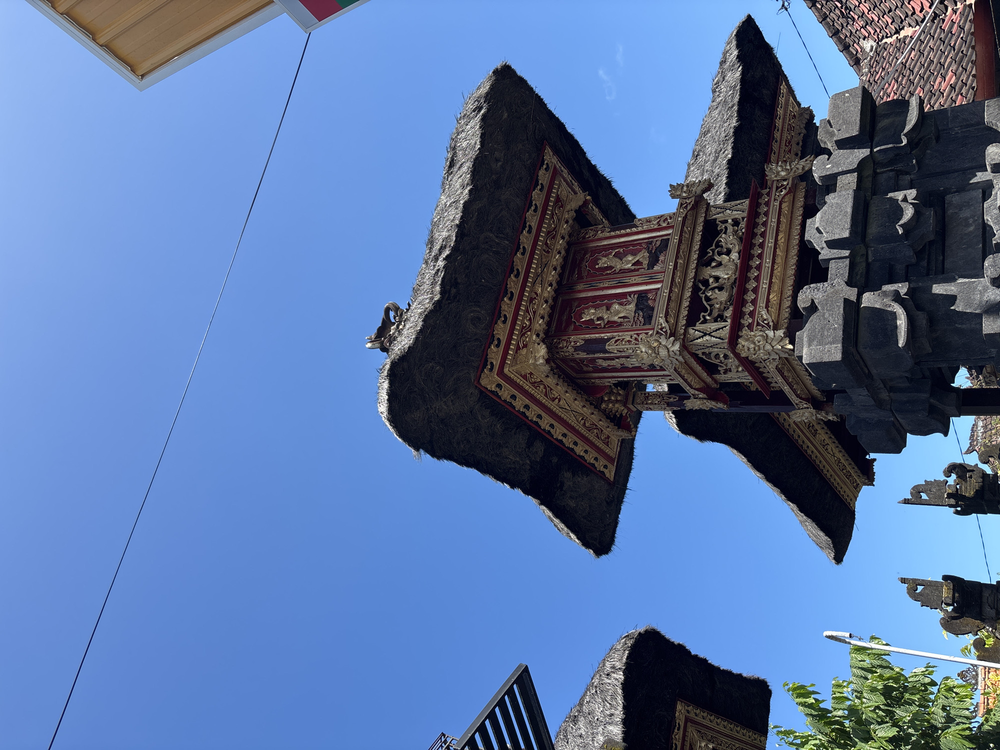 The low-angle shot depicts a traditional Balinese structure against a clear blue sky in Kuta Selatan, Indonesia. The structure features a multi-tiered roof with dark, thatched coverings and elaborate ornate carvings in red and gold. To the right, the structure incorporates dark stone elements with intricate detailing. A sign, partly visible in the upper left corner, suggests a commercial establishment (7-Eleven). At the bottom right, a leafy tree is partially visible, obscuring a street lamp. Other parts of similar Balinese structures are seen at the bottom, also against the sky. A thin black wire stretches across the upper part of the image.
