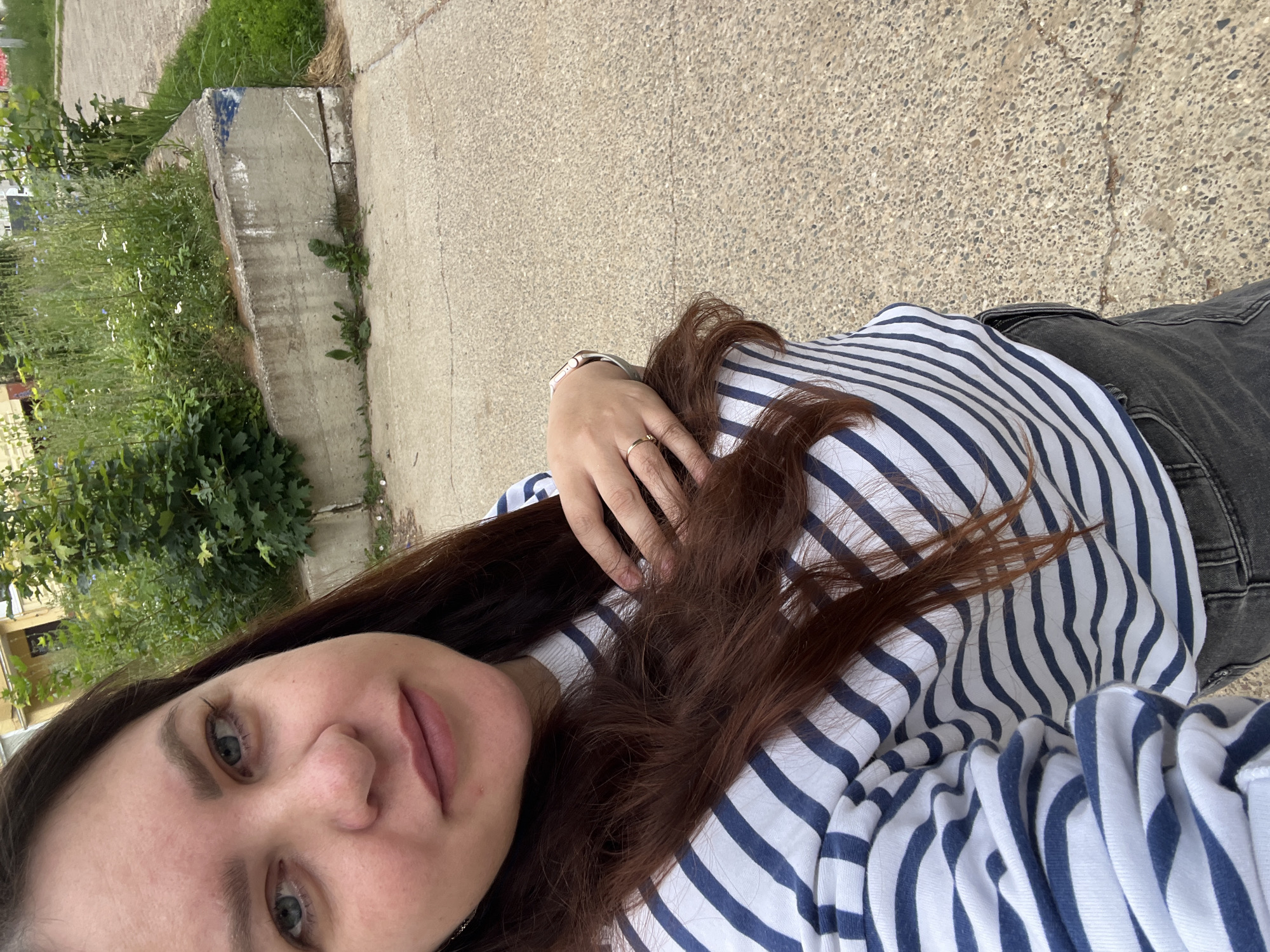 A selfie is taken outdoors, with the subject's face and upper body filling the left half of the frame. The subject has fair skin, light blue eyes, and long, dark brown hair. They are wearing a striped long-sleeved shirt with navy blue and white stripes, and dark gray jeans. A hand with rings is resting on their hair. The background features a concrete retaining wall with greenery and some plants growing on top. Beyond that, a paved surface is visible, likely a sidewalk or path, and a building with yellow siding can be seen in the distance. The overall setting appears to be an urban or suburban environment.