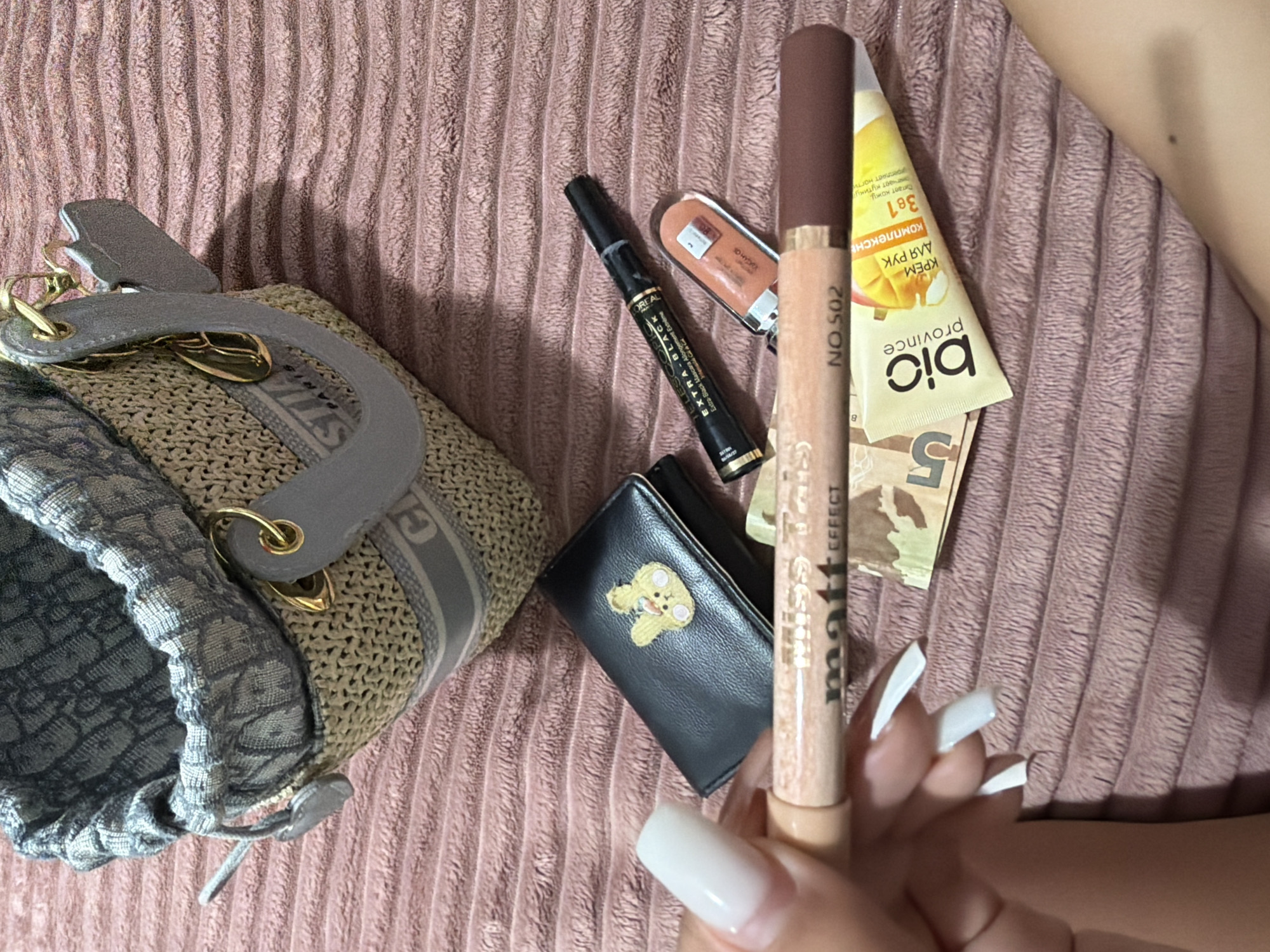 The image shows a collection of cosmetic items and a small wallet on a pink, textured surface, possibly a bedspread. In the background, a portion of a straw-like bag with gray accents and a Dior logo is visible. The items in the foreground include a black wallet with a small embroidered yellow bunny, a black mascara tube labeled "L'OREAL TELESCOPIC EXTRA-BLACK", a tube of lip gloss or balm, a makeup pencil labeled "miss tati matt effect NO.502", and a yellow tube of hand cream labeled "bio province" with Cyrillic text. There are also two Ukrainian 5 hryvnia banknotes partially visible. A person's hand with white, almond-shaped fingernails is holding the makeup pencil. The scene is well-lit, with the subjects in focus. There is no indication of the location being in Digah, Azerbaijan.