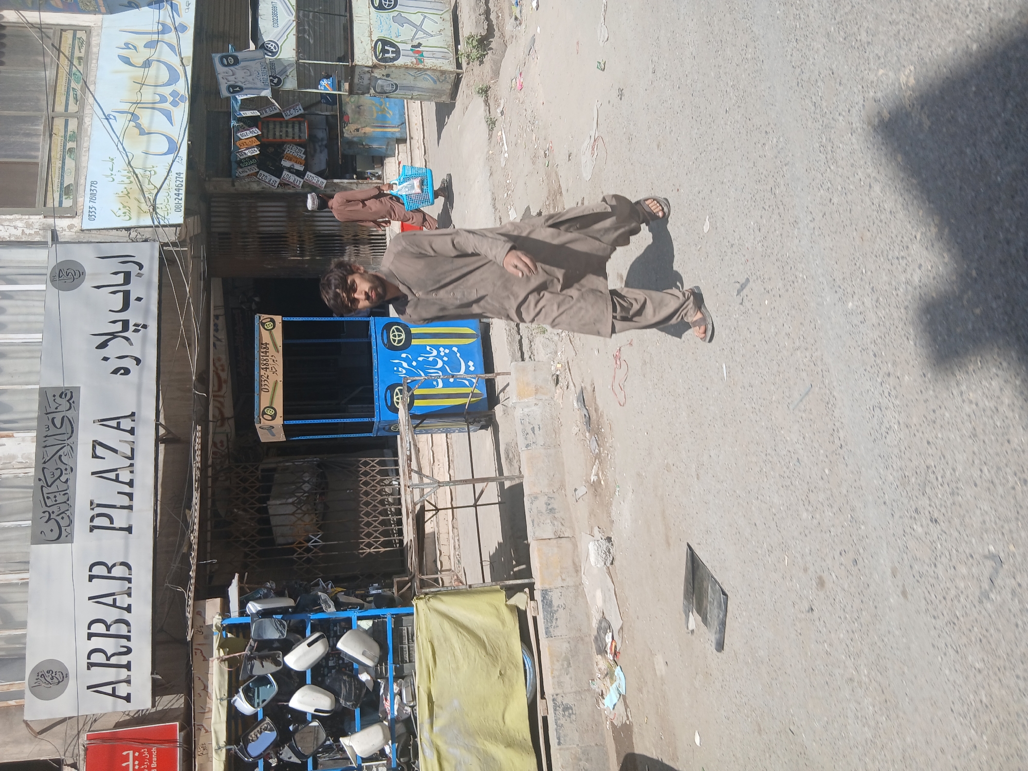 The image shows a street scene in Quetta City Tehsil, Pakistan. A man wearing a shalwar kameez walks along the side of the road. Behind him, a second person wearing a similar outfit is walking in the same direction. The storefront "ARBAB PLAZA" is visible on the left of the frame, displaying Pakistani and Urdu writing. Another business is visible above it. There are various car parts and accessories on display, including mirrors. The road appears dusty and littered. There is also a vehicle with blue siding with yellow stripes. A series of license plates is hanging next to it.