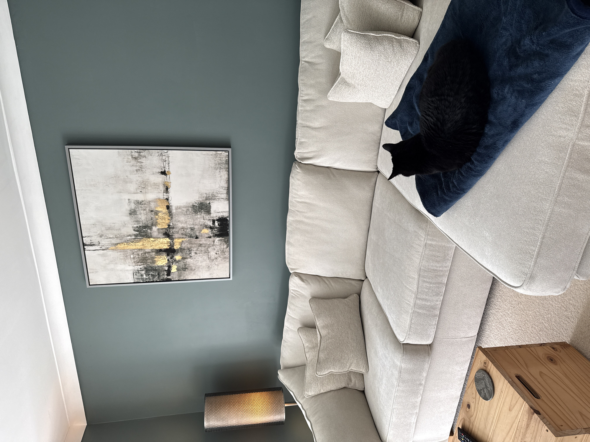 A room interior is depicted in this top-down view. The main features are a light-colored sectional sofa with a navy blue blanket, and a black cat lying on the blanket. Two textured pillows of the same color as the sofa are placed on it. A framed abstract painting hangs on the wall. Below it, partially visible, is a lamp with a textured lampshade. The wall is painted in a teal-grey color. To the right of the sofa, there is a light-colored wooden crate. The location in East Suffolk, United Kingdom cannot be determined based on this image.