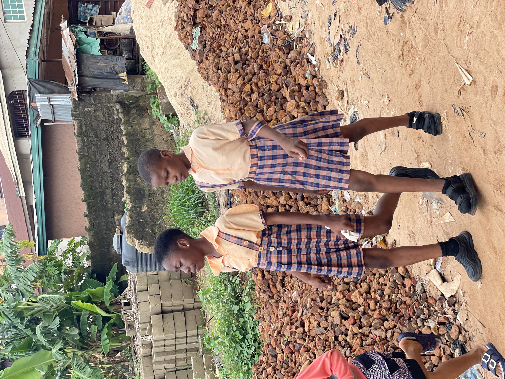 Two children in school uniforms stand on a pile of reddish-brown rocks or stones in Onitsha, Nigeria. They are wearing black shoes. The location appears to be an outdoor area with sand, and scattered debris or trash. In the background, there are buildings, plants, and stacks of building materials.
