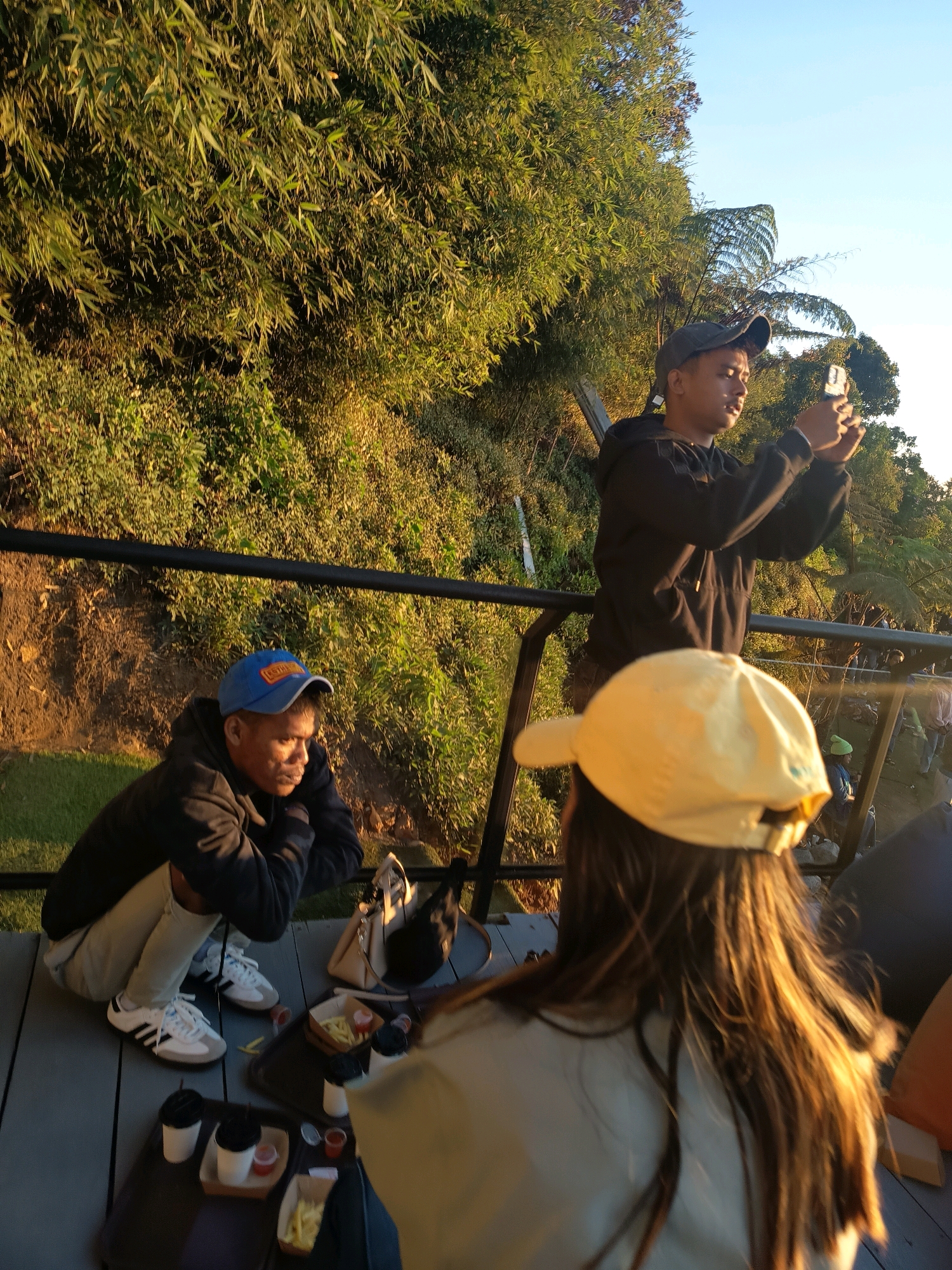 In Keditan, Indonesia, at an outdoor viewpoint during golden hour, three individuals are present. One man, wearing a black hoodie and a blue baseball cap, is crouched on a wooden deck, looking down and to the side. Beside him, a second man in a black hoodie and a dark baseball cap stands and is using a smartphone to take a picture. In the foreground, a third person, facing away from the camera and wearing a yellow baseball cap, is seated. The scene is framed by lush green foliage and a clear sky. On the deck, trays of food, including french fries and drinks, are visible.