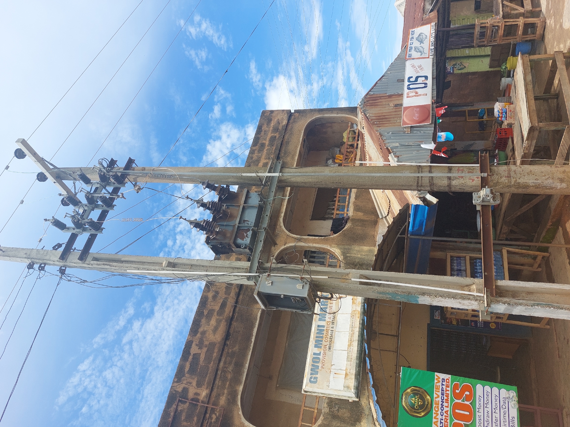 This is a street-level, eye-level medium shot of a commercial area in Barakin, Nigeria. A large, weathered utility pole with a transformer and numerous wires dominates the center of the frame. The pole is situated in front of a two-story building with a textured, earthy-brown facade. The building has arched openings and balconies.
To the left of the utility pole, a sign reads "GWOL MINI MART PROVISION, COSMETICS & WINE ENJOY (WHOLESALE & RETAILS)". Below this sign, another banner displays "ORANGEVIEW MULTI-CONCEPTS NIGERIA LIMITED. POS Deposit Money, Withdraw Money, Transfer Money, Airtime/Data, Bills".
To the right of the utility pole, a corrugated metal roof covers a storefront. A sign above this storefront reads "POS AVAILABLE HERE" with the Coca-Cola logo to its left. Further to the right, another sign depicts three fish and reads "BUY YOUR ICE FISH HERE LIVE AND LETS LIVE". Several wooden tables and chairs are scattered around the area, some with goods on them, suggesting a marketplace or informal retail setting. In the background, there are more storefronts and various objects, including bottles of water. The sky is blue with scattered white clouds. No people are visible in the image.