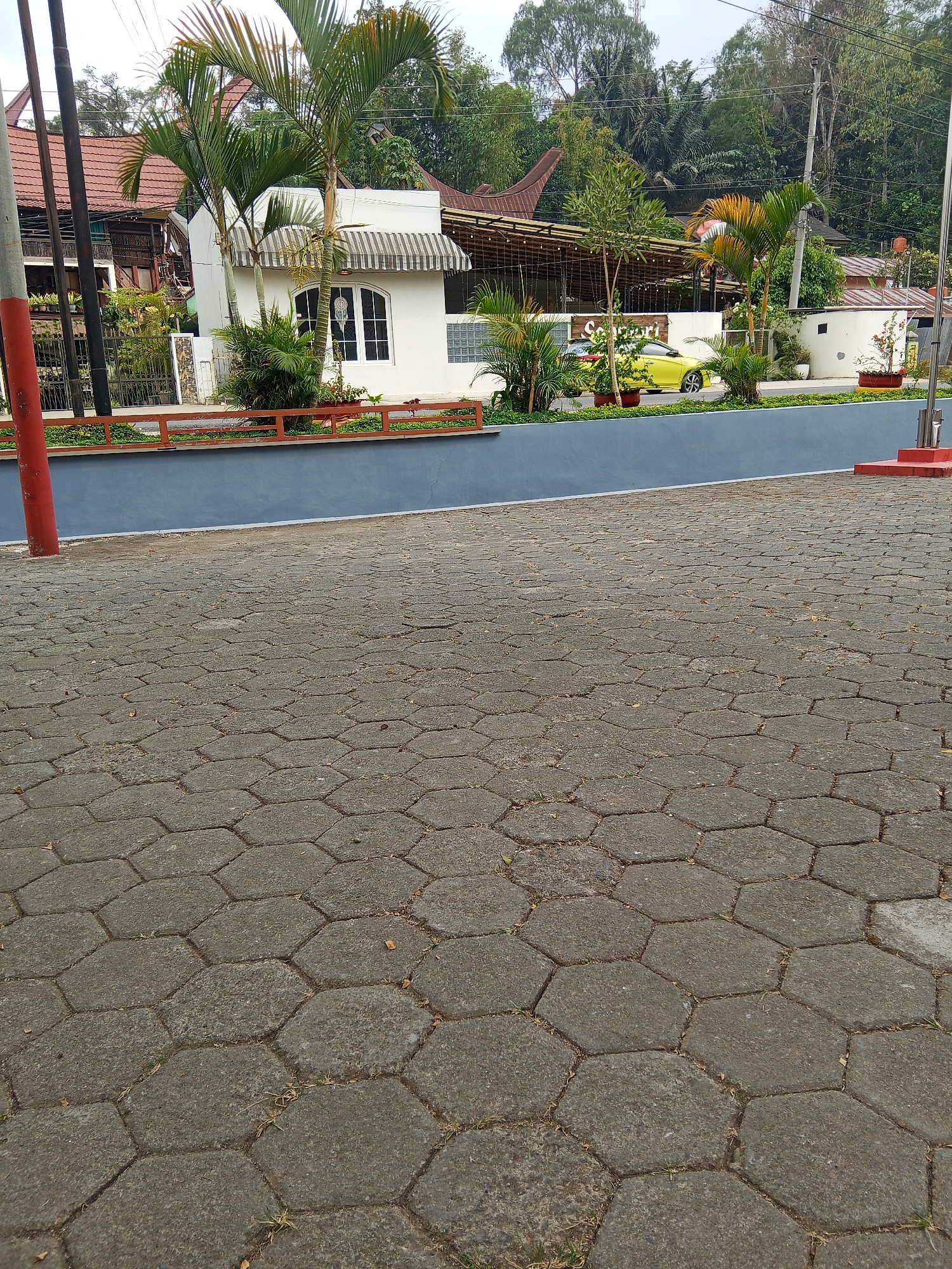 The image depicts a street scene in Bungin, Indonesia, dominated by a hexagonal paver stone ground in the foreground. In the background, a white building with a striped awning over a window is visible. Adjacent to it, a building with a covered outdoor seating area, illuminated by string lights, is present. A yellow car is parked in front of this structure. The scene is bordered by a blue wall and a reddish-brown railing with planters. Green vegetation, including palm trees, is scattered throughout the background, with a wooded hillside in the distance. Power lines and poles are also visible. The overall impression is of a commercial or hospitality area, possibly a cafe or restaurant, with a paved outdoor space.