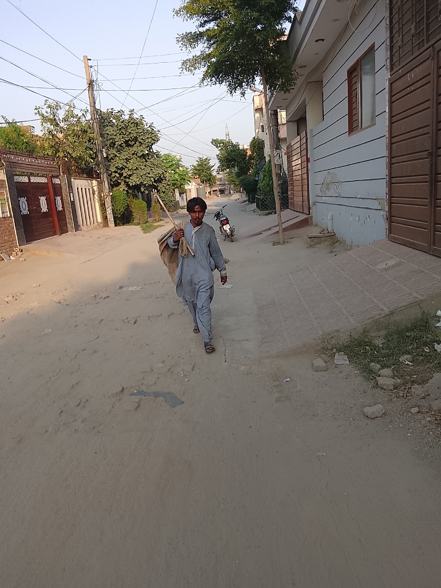 A man walks down a dirt road in Unknown, Unknown. He wears a light grey shalwar kameez and sandals. He carries a stick over his shoulder with a burlap sack attached to it. To the right of the man are buildings with light grey siding and a brown garage door. There is a small patch of grass next to the building. Across the street, there are trees and houses with closed brown doors. A motorcycle is parked between the man and the houses across the street. Power lines run above the street.