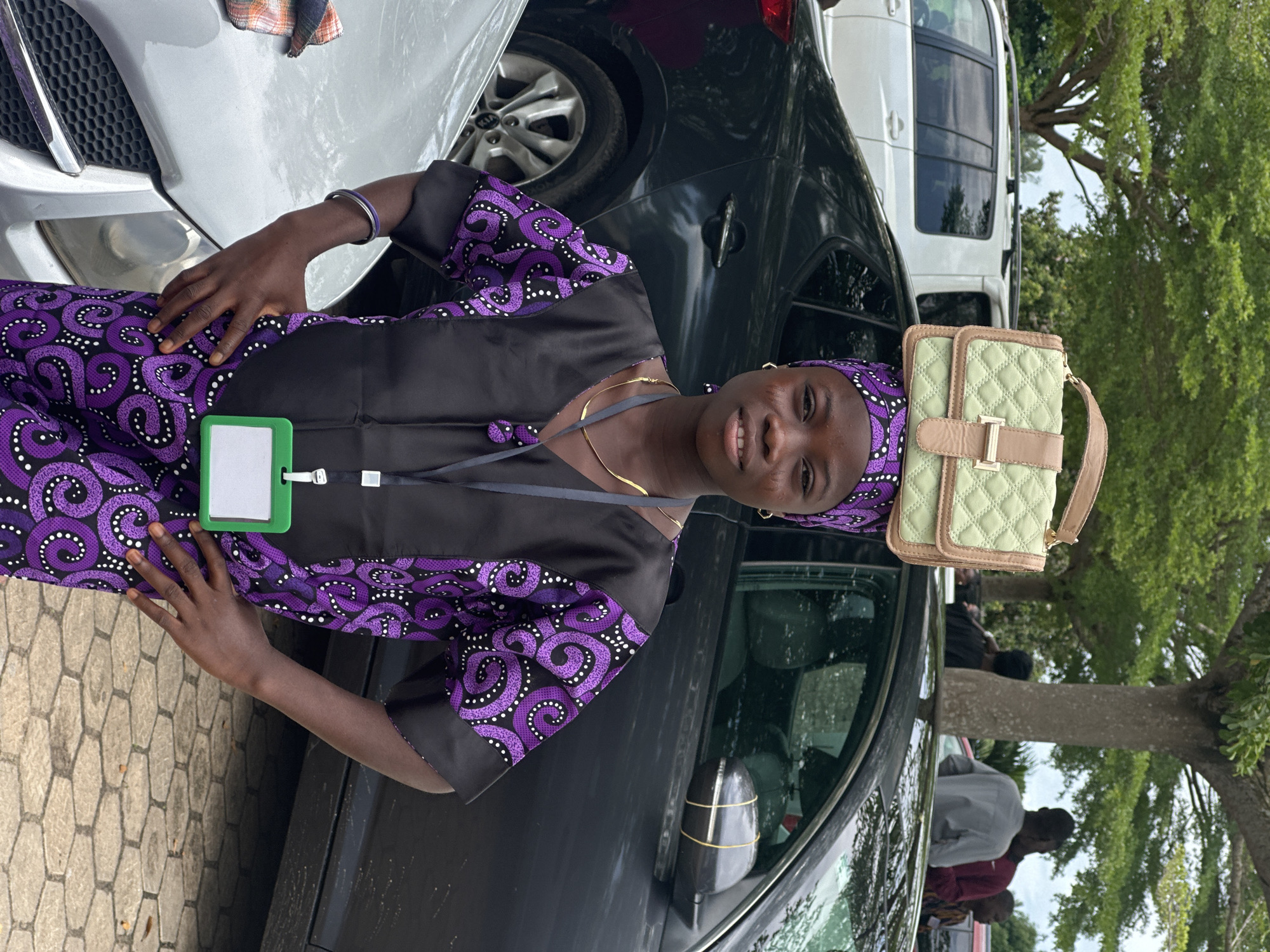 In Itoga, Nigeria, a young girl stands in front of a dark-colored sedan with her hands on her hips. She wears a black and purple patterned traditional outfit and a matching headscarf. A light green quilted handbag rests on top of her head. A green lanyard with an ID badge hangs around her neck. To her left, a silver car is partially visible. In the background, trees and other vehicles, including a white SUV, are present.