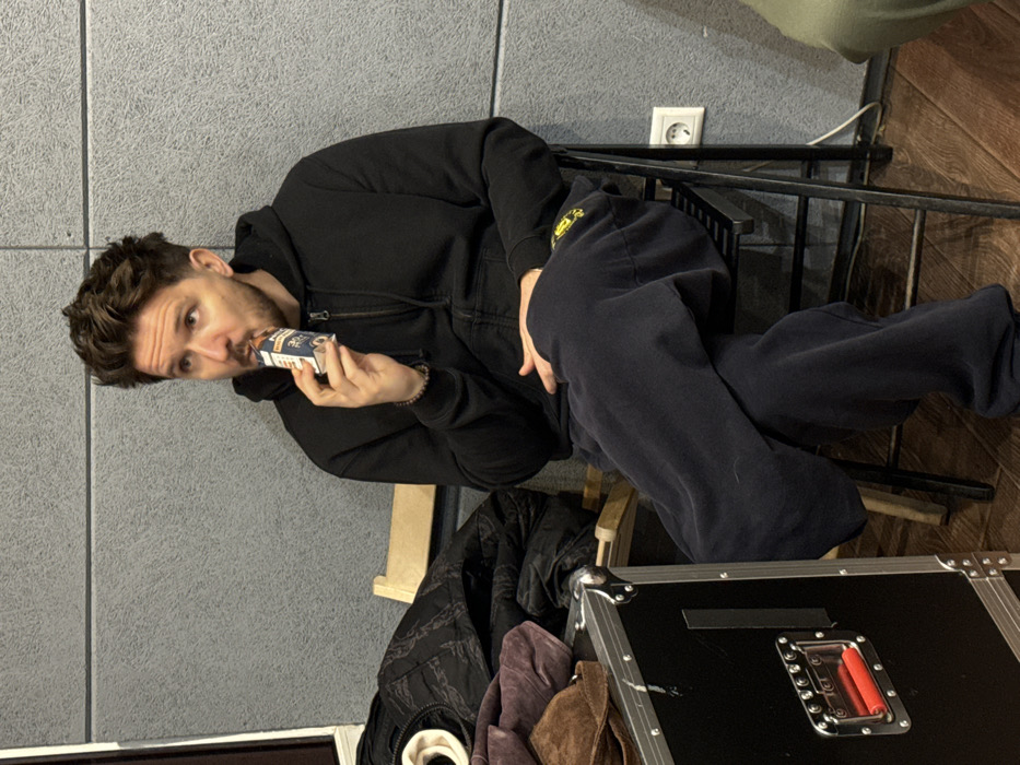 An adult male is seated on a black metal folding chair within an interior space in Moscow, Russia, consistent with a recording studio or rehearsal room. The subject, exhibiting short, wavy brown hair and light facial stubble, is dressed in a black zippered hoodie and dark blue sweatpants. A dark beaded bracelet is visible on his right wrist. He is actively consuming a beverage from a small, light-colored rectangular carton via a straw, holding the carton to his mouth. His gaze is directed upwards and to the left of the frame. The immediate environment features a textured gray wall, consistent with acoustic paneling, above a wooden floor. A large black equipment flight case with silver trim and a red handle is positioned to the left. Other visible objects include a brown hat, dark folded clothing, and a partial view of a light-colored wooden chair. A white two-socket European-style power outlet with an attached white cable is mounted on the gray wall to the right.