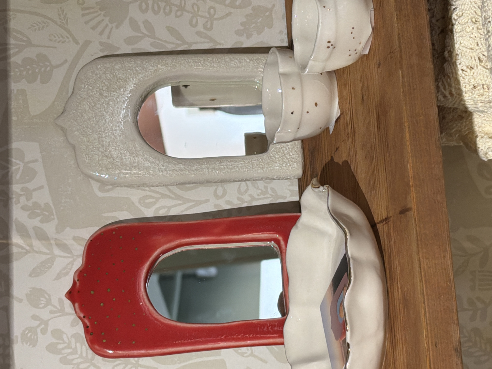 The image depicts a close-up, eye-level view of a wooden shelf containing decorative ceramic items. On the left, a red, arched mirror with small green dots is displayed. To its right is a cream-colored, arched mirror with a crackled glaze. In front of these mirrors are two small, white ceramic bowls with small dark dots. The shelf itself is made of dark wood, and a patterned wallpaper with floral motifs serves as the background. The scene is well-lit, suggesting an indoor setting, possibly a shop or a home display. The image does not provide any information about the city of Suzdal, Russia.