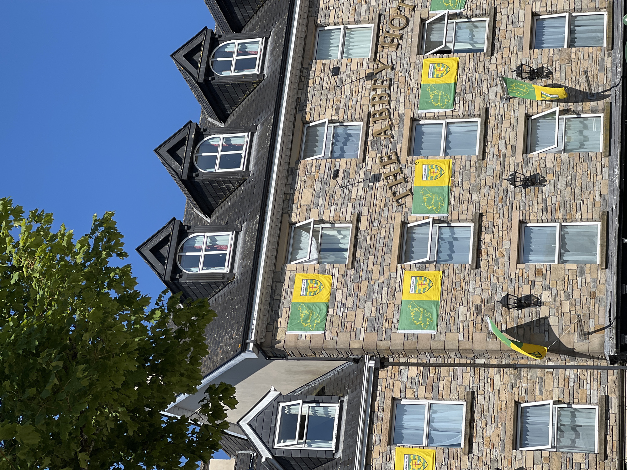 The exterior of The Abbey Hotel in Donegal Town, Ireland, is shown on a clear, sunny day. The stone facade of the building is decorated with multiple flags, predominantly green and yellow, bearing the crest of Donegal GAA. These flags are hung from the windows and walls of the hotel, some displaying the text "DÚN NA NGALL" and "Donegal". A large tree with green leaves partially obscures the upper left portion of the building. The hotel has multiple floors with numerous windows, some of which are open. The hotel's name, "THE ABBEY HOTEL", is visible in gold lettering above the flags. Several decorative black lanterns are mounted on the facade. The sky is a vibrant blue.