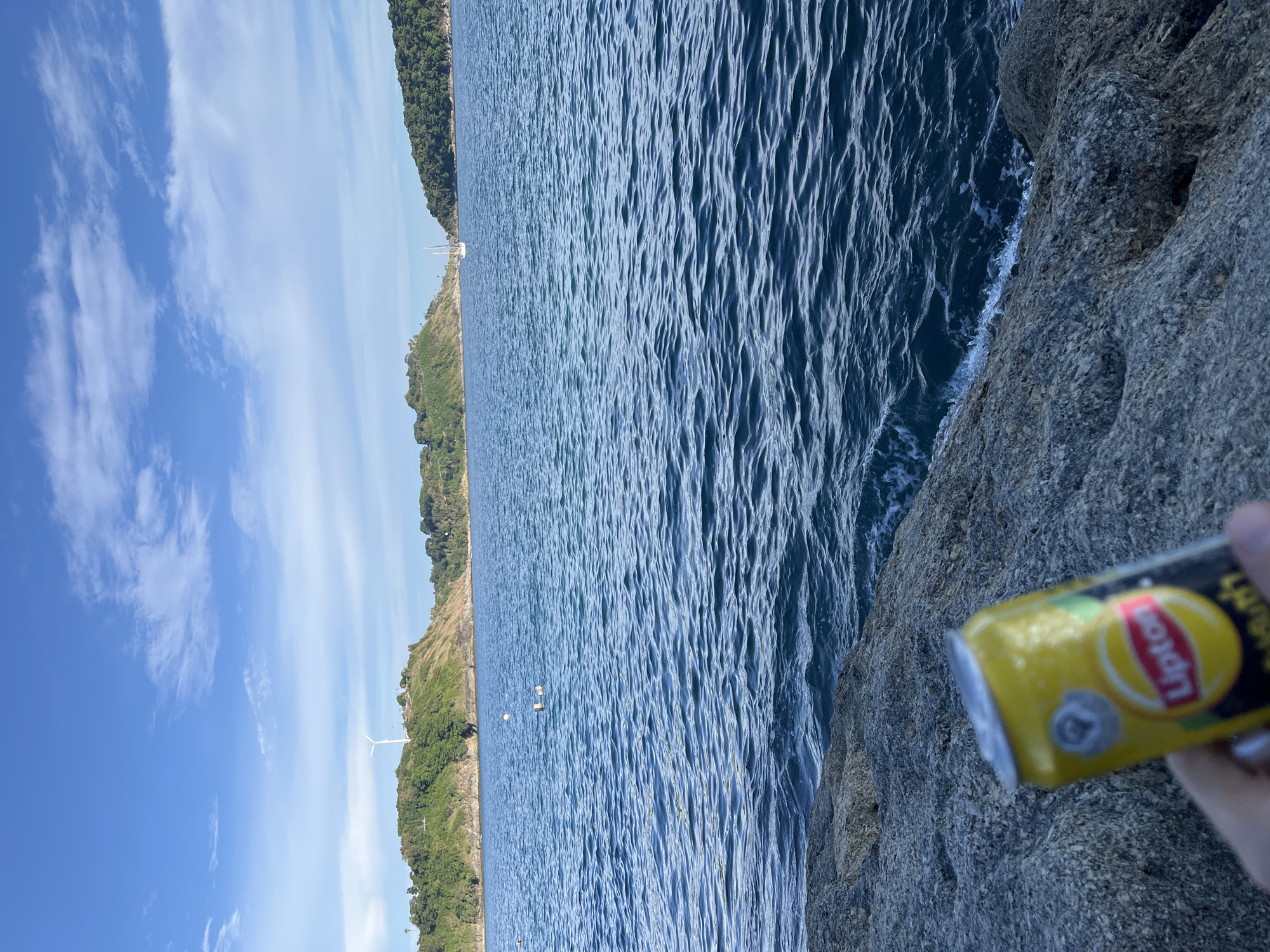 A yellow Lipton Iced Tea can is held in the foreground, in focus, by a person's hand which is partially visible. The can is positioned on a rocky shore, with the dark blue ocean stretching out behind it. The water is rippled and shows some small waves crashing against the rocks. In the distance, two tree-covered headlands frame a bay, with a white sailboat visible in the center. A single wind turbine stands atop the headland to the left. The sky is blue with scattered white clouds. The scene appears to be located in Rawai, Thailand.