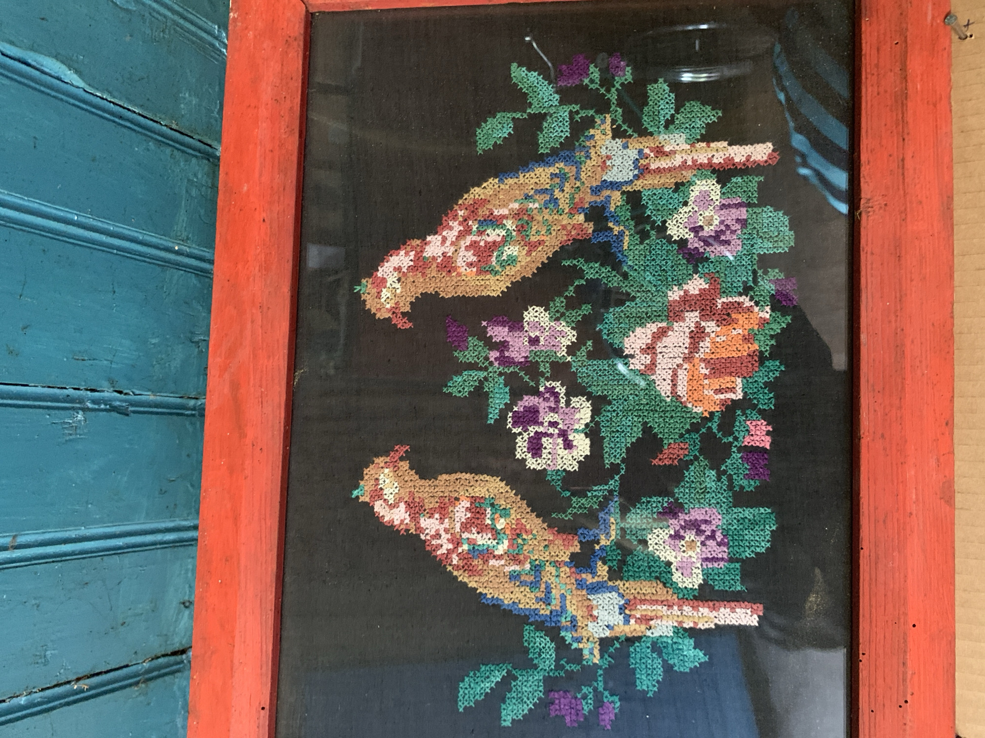 The image shows a framed cross-stitch artwork featuring two colorful parrots perched on a branch with flowers. The parrots are depicted in shades of orange, yellow, blue, and red, with intricate patterns on their feathers. They are facing each other, with lush green leaves and purple and pink flowers surrounding them. The background of the cross-stitch is black. The artwork is enclosed in a rustic red wooden frame. The frame is placed against a textured, teal-painted wooden surface, likely a wall or ceiling, which is composed of planks. The lighting suggests an indoor setting, possibly a home or workshop in Відзаўскі сельскі Савет, Belarus.