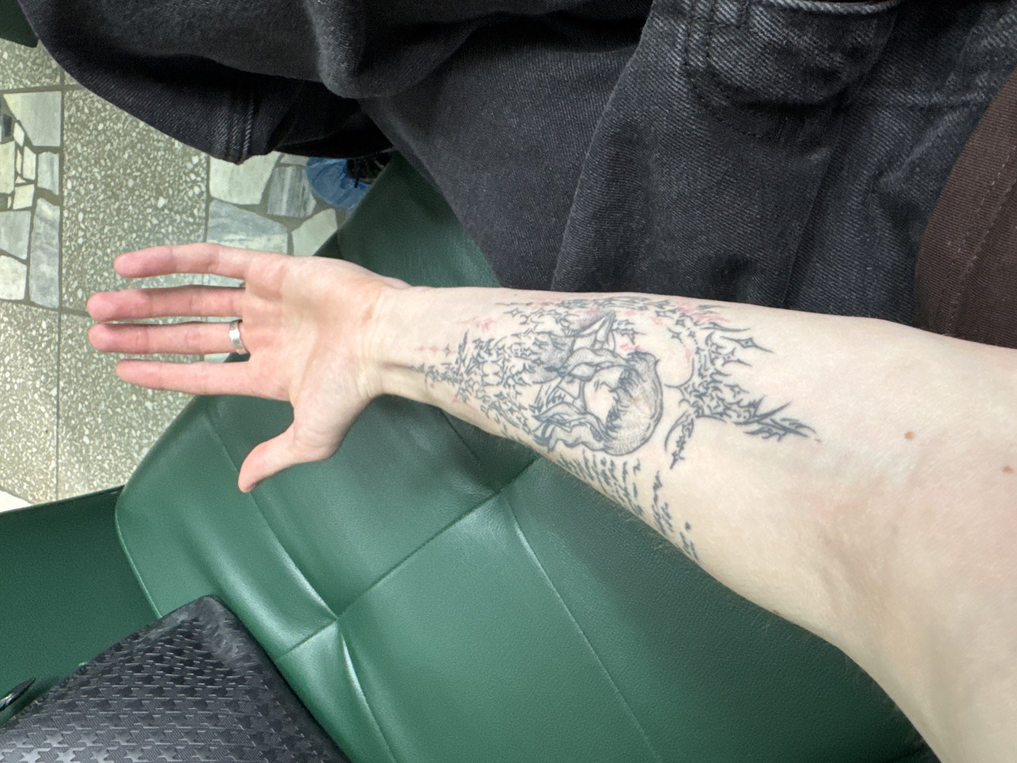 A photograph taken indoors shows a fair-skinned person's arm extending into the frame, with a prominent tattoo of a jellyfish-like figure on the forearm. The person is wearing a silver ring on their left ring finger. The arm rests on a green, cushioned surface, possibly a chair or seat. In the background, there's a glimpse of a dark, textured item that could be a bag or clothing, and above that, part of what appears to be dark-colored denim clothing. The location of the photograph is reported to be Chaikovsky, Russia, but there are no location details that can be verified by visual features of the image.