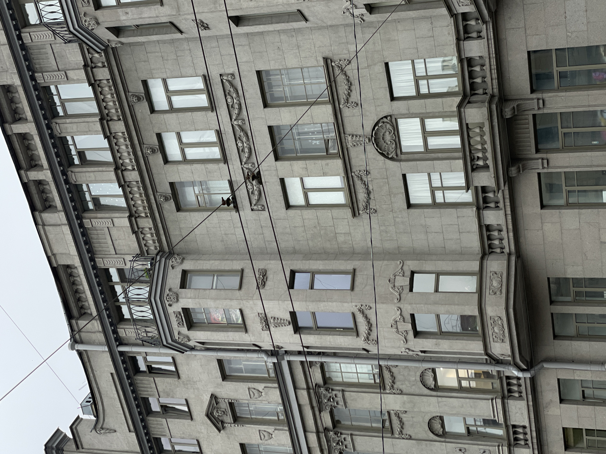 The image depicts a multi-story building in Saint Petersburg, Russia. The building's facade is gray and features numerous windows and ornate architectural details, including decorative moldings and small balconies. Power lines run across the front of the building. The sky is overcast.