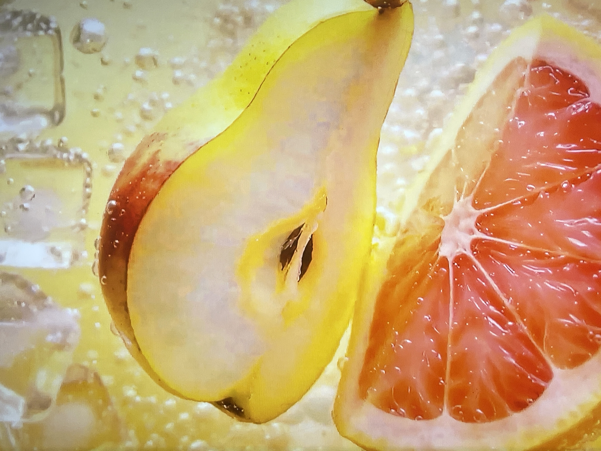 The image shows a close-up, macro shot of a sliced pear and a slice of grapefruit submerged in a bubbly, yellow liquid, possibly a drink or soda. Ice cubes are visible in the background on the left side of the image. The pear is cut lengthwise, revealing its seeds. The grapefruit slice showcases its segmented flesh and juicy texture. The lighting highlights the water droplets and bubbles clinging to the fruit and ice. There are no people or specific landmarks present in the image. The scene is aquatic and food-focused. There is no information to suggest the location in the city of Shakhovskaya, Russia.