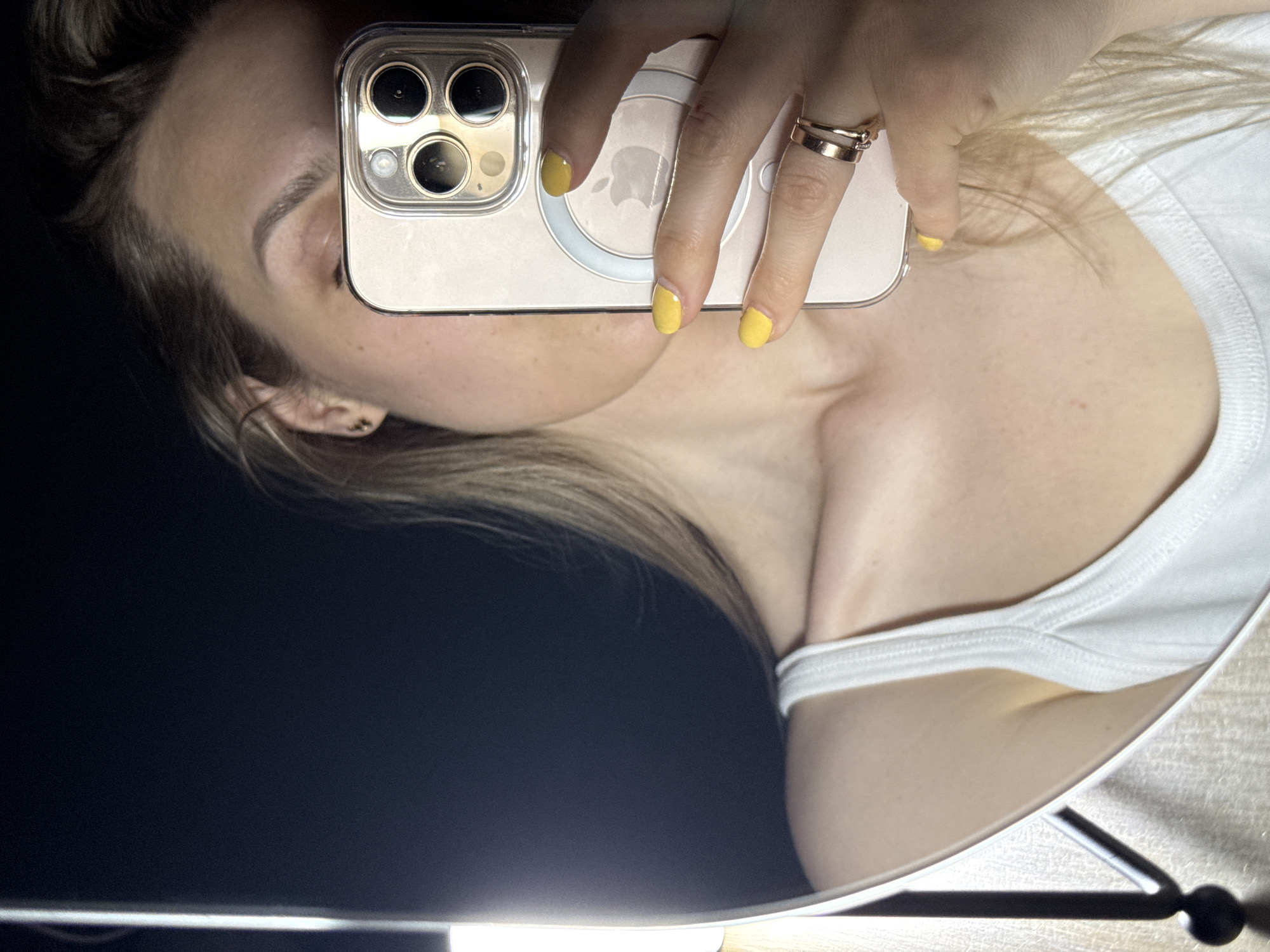 A rotated mirror selfie shows a person, likely female, capturing their reflection. The person's head is on the left side of the frame, with her right eye, eyebrow, and part of her forehead visible. Her light brown hair is loosely styled. She holds a smartphone, appearing to be an iPhone with three camera lenses and a MagSafe ring with a faint Apple logo, in her right hand. The phone, encased in a clear cover, largely obscures her face. Her right hand features bright yellow-painted fingernails and two gold-toned rings on her ring finger. She wears a white tank top, showing her neck, shoulder, and upper chest. The selfie is taken in a reflective surface, possibly a mirror or a dark, polished table. A bright, rectangular light source is strongly reflected at the bottom of the frame. The background behind the person is dark. The scene depicts a casual, indoor moment, likely in a dimly lit room, focusing on a self-portrait. No specific location cues for Chaikovsky, Russia, are discernible within the image.
