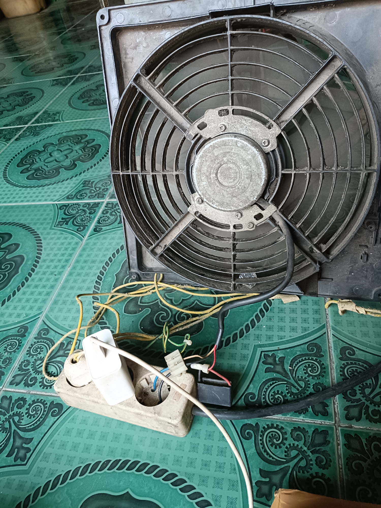 A dusty, circular fan with a metal grill is positioned on a green tiled floor. Wires and a white power strip with a plugged-in adapter are connected to the fan, suggesting it is either being repaired or has been disassembled. The green tiles have an intricate, repeating pattern. The scene appears to be indoors, with no visible windows or doors. The location is described as Telagamurni, Indonesia, though no specific urban landmarks are visible.